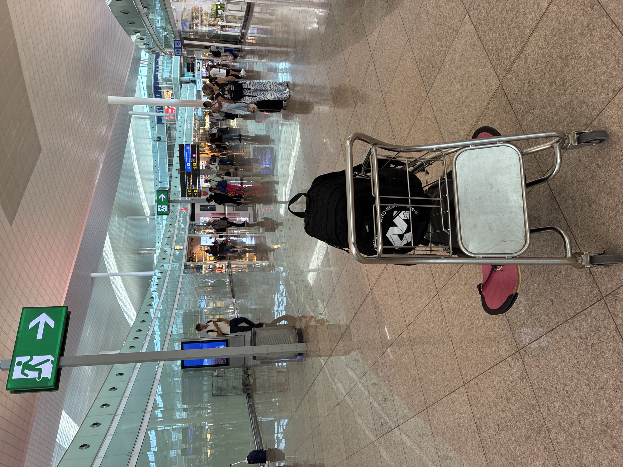 This image depicts the interior of an airport terminal, likely at Josep Tarradellas Barcelona–El Prat Airport in El Prat de Llobregat, Spain, given the visual cues of an international airport environment. In the foreground, a luggage cart is positioned on a polished tiled floor, with a black backpack placed on top. A pink item, possibly a sandal or mat, is visible beneath the cart. The background reveals a busy airport concourse with numerous people walking, some with luggage. Several shops and information signs are visible along the walls, which are lined with reflective glass panels. An illuminated green exit sign with an arrow pointing to the right is prominently displayed on a pole, indicating an emergency exit. The ceiling is made of white modular panels, and large columns support the structure. The overall impression is one of a modern, well-lit, and active airport transportation hub.