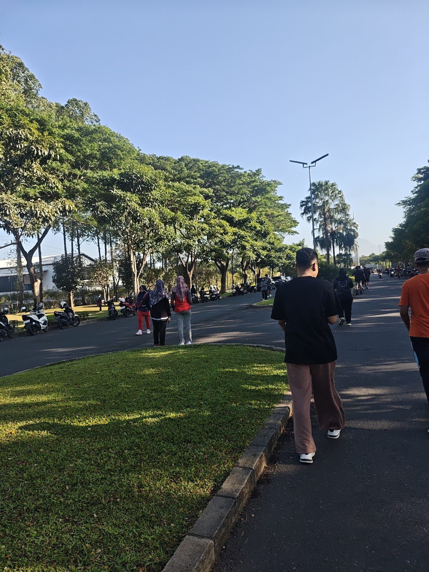 The image depicts an outdoor scene in Bacin, Indonesia, likely a park or a recreational area. The sky is clear and blue. In the foreground, a man wearing a black t-shirt and brown pants is walking on an asphalt path, facing away from the camera. To his left, a well-maintained grassy area with dappled sunlight runs alongside the path, bordered by concrete curbs.
Further down the path, several people are walking or standing. Two women in the mid-ground are wearing headscarves and athletic wear, one in red and the other in a greyish-blue outfit. Behind them, more individuals are visible, also dressed casually for outdoor activity. On the left side of the image, parked motorcycles are lined up along the road.
The background is filled with lush green trees, characteristic of the tropical climate of Indonesia. A street lamp with two angled lights is visible in the mid-background. Beyond the trees, buildings and what appears to be a distant mountain range can be seen, suggesting a semi-urban or suburban setting. The overall atmosphere is one of a bright, sunny day with people engaged in recreational activities.