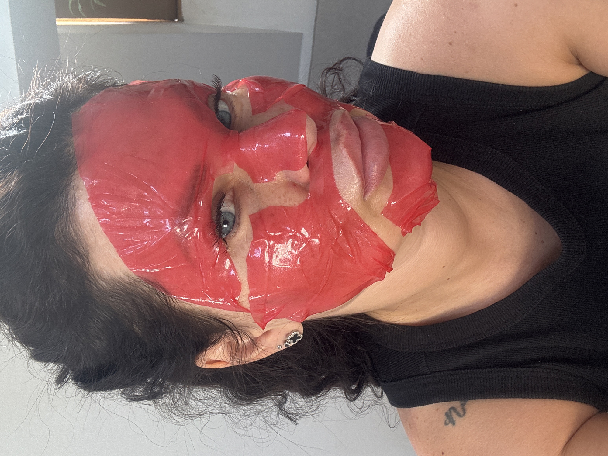 A woman with dark hair pulled back is wearing a red sheet mask that covers most of her face, obscuring her forehead, nose, and cheeks. Her eyes are visible and are blue. She is wearing a black tank top and a small, silver earring shaped like a four-leaf clover on her left ear. A tattoo is visible on her left shoulder. The background is a light-colored wall, and a portion of a window with blinds is visible on the right. The lighting suggests an indoor environment. There is no discernible information in the image that places it in Katowice, Poland.