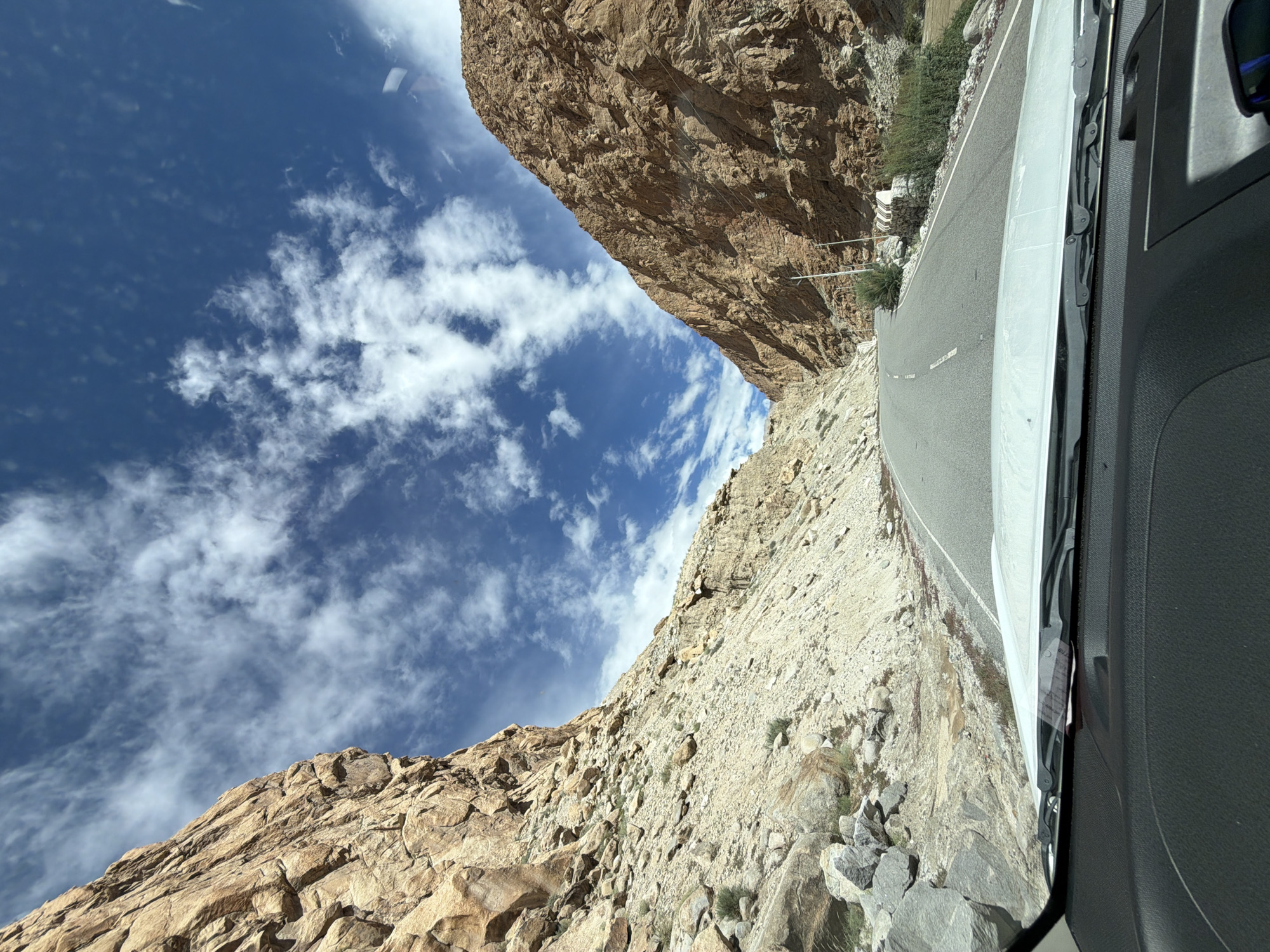 The image depicts a view from inside a vehicle, looking out through the windshield at a road winding through a rocky canyon. The canyon walls are steep and made of tan and brown rock, with scree slopes leading down to the road. The sky is a vibrant blue with scattered white clouds. Sparse green vegetation is visible along the sides of the road and on the lower slopes. The road itself is paved and has white dashed lines indicating lanes. There are no people or animals visible in the image. The location is stated to be in the city of Kungyamdo, India, which is a mountainous region.