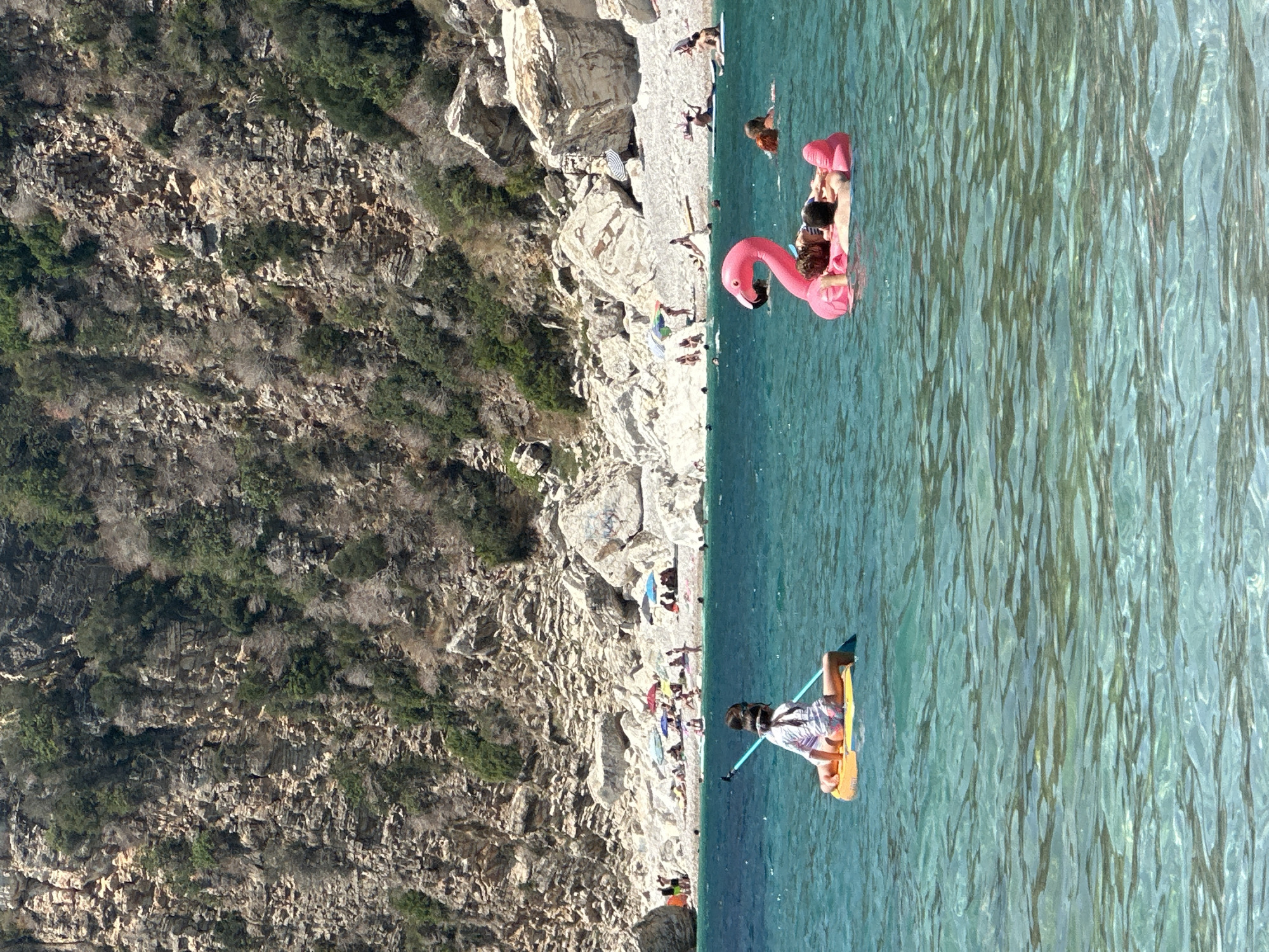 The image depicts a vibrant beach scene in Mirište, Montenegro, under a clear, bright sky. The foreground is dominated by the sparkling, turquoise waters of the Adriatic Sea, with clear visibility down to the sandy bottom in shallower areas. In the middle ground, two individuals are enjoying the water. On the left, a person is seated on a yellow paddleboard, holding a blue paddle. Further to the right, two people are lounging on a large, inflatable pink flamingo float.
The background features a pebbly beach crowded with sunbathers, beach umbrellas, and some rocky outcrops. A steep, rocky cliff face, covered in sparse vegetation, forms the backdrop to the beach. The overall atmosphere is one of a serene and sun-drenched summer day at a popular coastal destination.