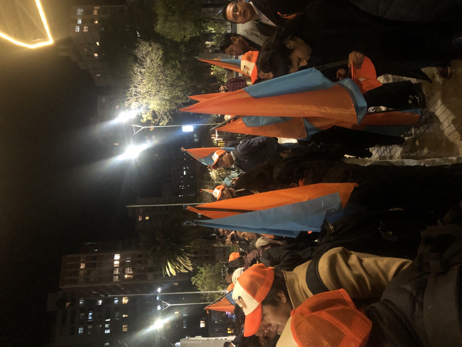 People with orange and white caps hold orange and blue flags in a public square at night. Streetlights illuminate the scene. Buildings rise in the background with lights visible in some windows. The group appears to be focused and attentive, possibly at a political gathering or demonstration.