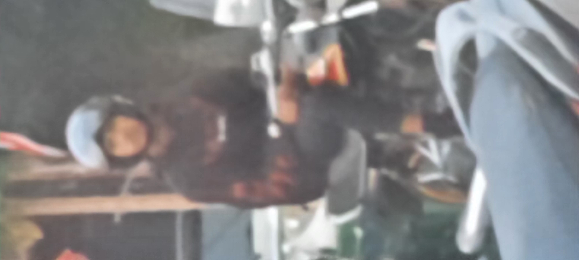 In Kanigoro, Indonesia, a person wearing a black hoodie and a motorcycle helmet with goggles is seated on a motorcycle. The person's face is partially obscured by the helmet. The background shows a blurred environment with foliage and what appear to be poles, suggesting an outdoor setting. The image is low-resolution and blurred, making it difficult to discern finer details. The motorcycle is dark-colored and appears to be an older model.
