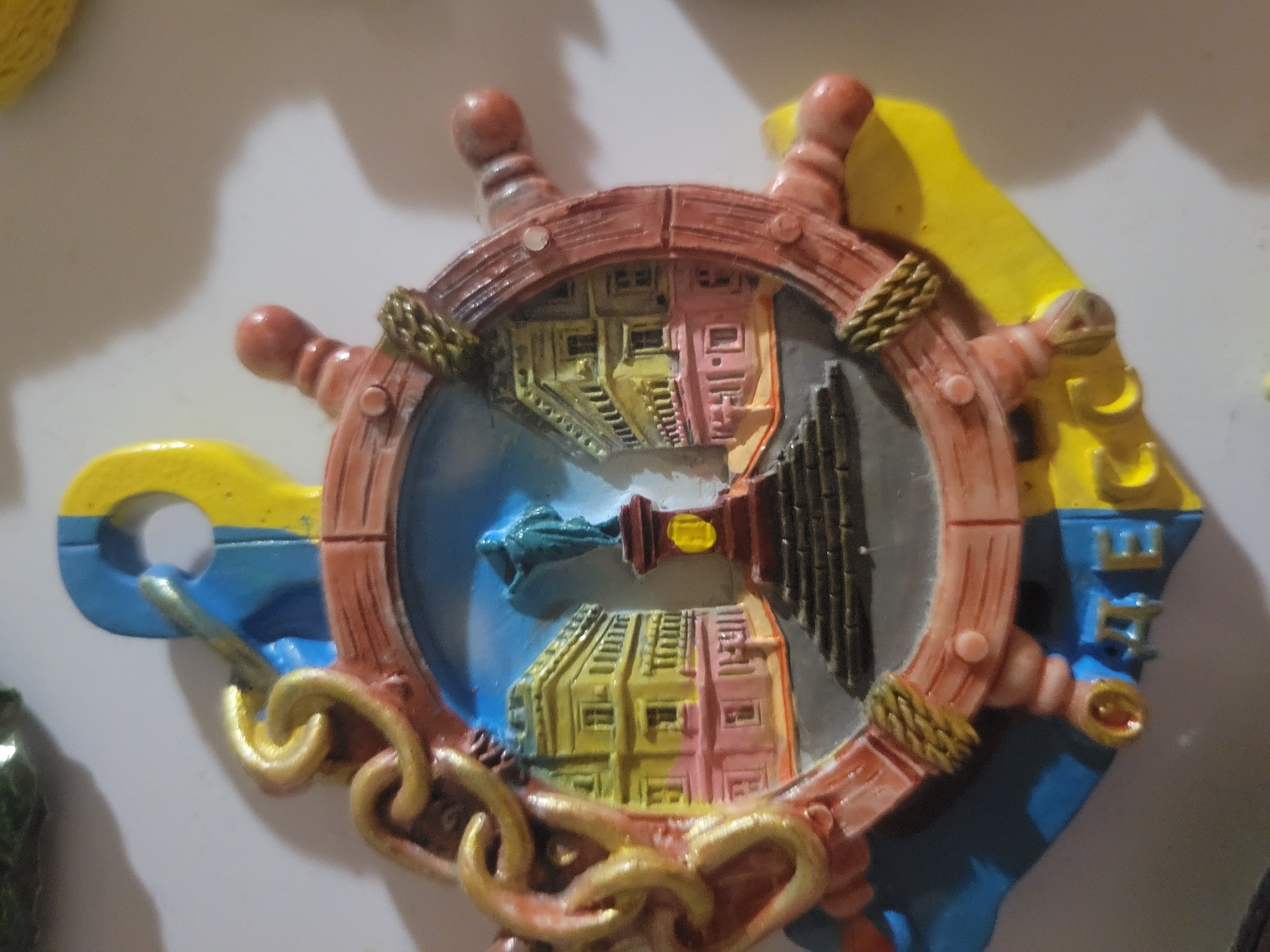 The image shows a souvenir magnet shaped like an anchor with a ship's wheel at its center. The ship's wheel depicts a scene with buildings and a statue in the middle. The magnet is predominantly blue and yellow, possibly representing the colors of the Belarusian flag. Below the ship's wheel, there are some Cyrillic letters visible, which are "ОДЕССА". This inscription indicates that the souvenir is related to the city of Odesa, Ukraine, not Belaazyorsk, Belarus. The magnet appears to be made of plastic or resin and is attached to a white surface.