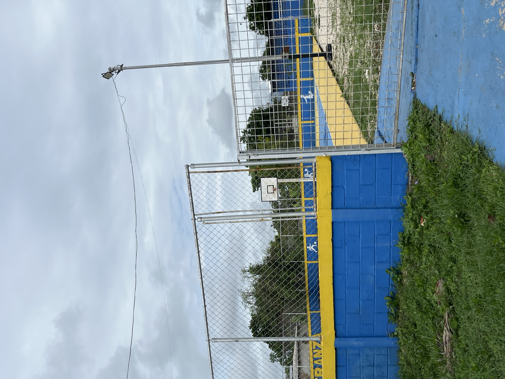 The image shows an outdoor recreational area, likely a sports court, in Tinaquillo, Venezuela. The court features a blue and yellow painted surface. A blue brick wall topped with a yellow band surrounds the perimeter. The word "ESPERANZA" is visible in white lettering on the yellow band. A chain-link fence encloses the area, with a metal gate in the foreground. A single basketball hoop is visible on the left side of the court. A lamppost with a spotlight is positioned behind the fence. The sky is overcast with clouds. There is grass and some dry foliage in the foreground.