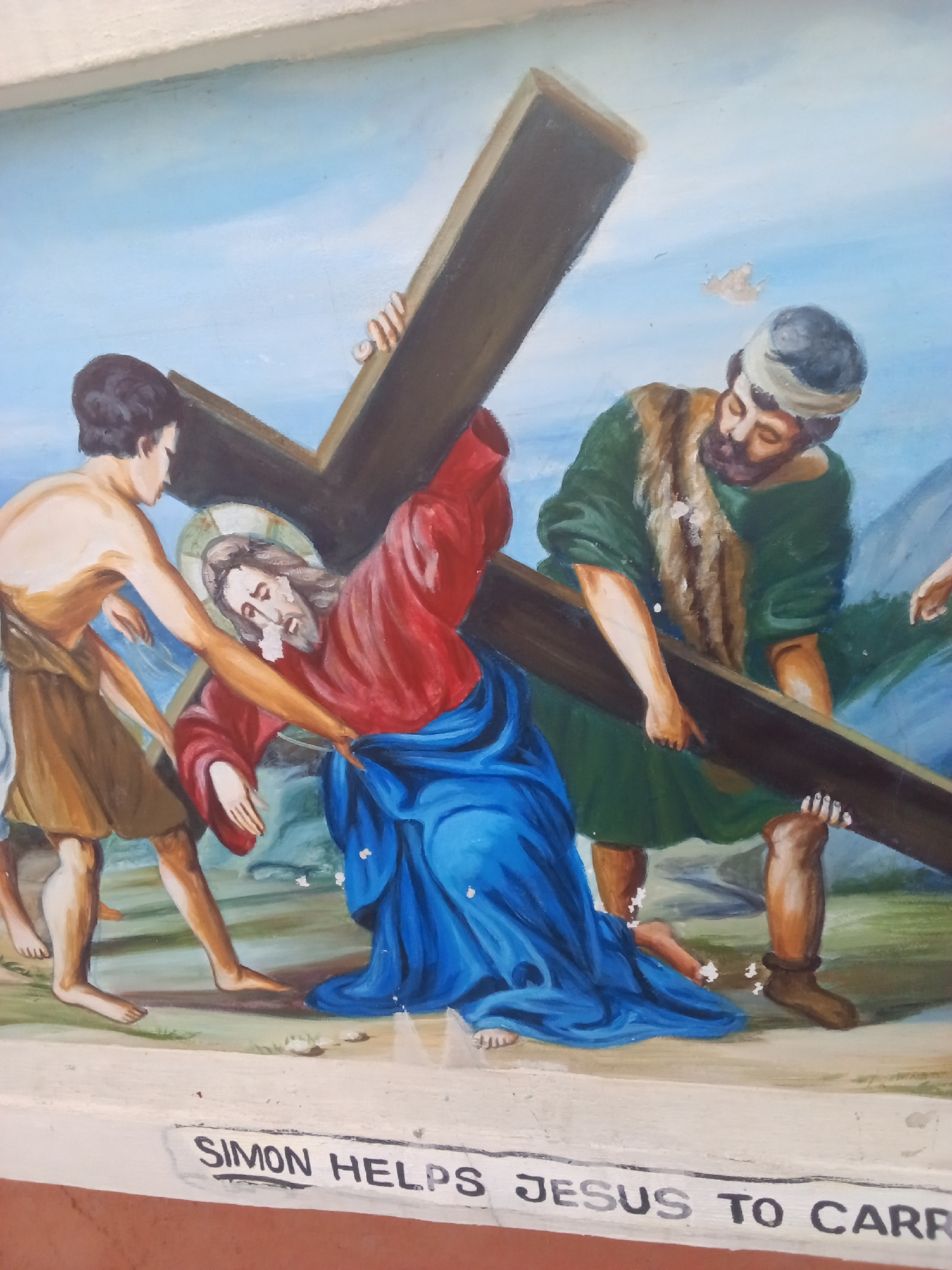 The image is a painting depicting the biblical scene of Simon of Cyrene helping Jesus carry the cross. Two men are assisting Jesus, who is struggling under the weight of a large wooden cross. One man is on the left, wearing a brown cloth. He is bare-chested and has dark hair. The other man is on the right, wearing a green tunic with fur, a head covering, and brown boots. Jesus is in the center, wearing a red robe and blue garment, with a halo above his head. The background shows a blue sky and vague landforms. A white dove flies in the upper left area. The image includes a caption stating "SIMON HELPS JESUS TO CARRY". The location is Ado, Nigeria.