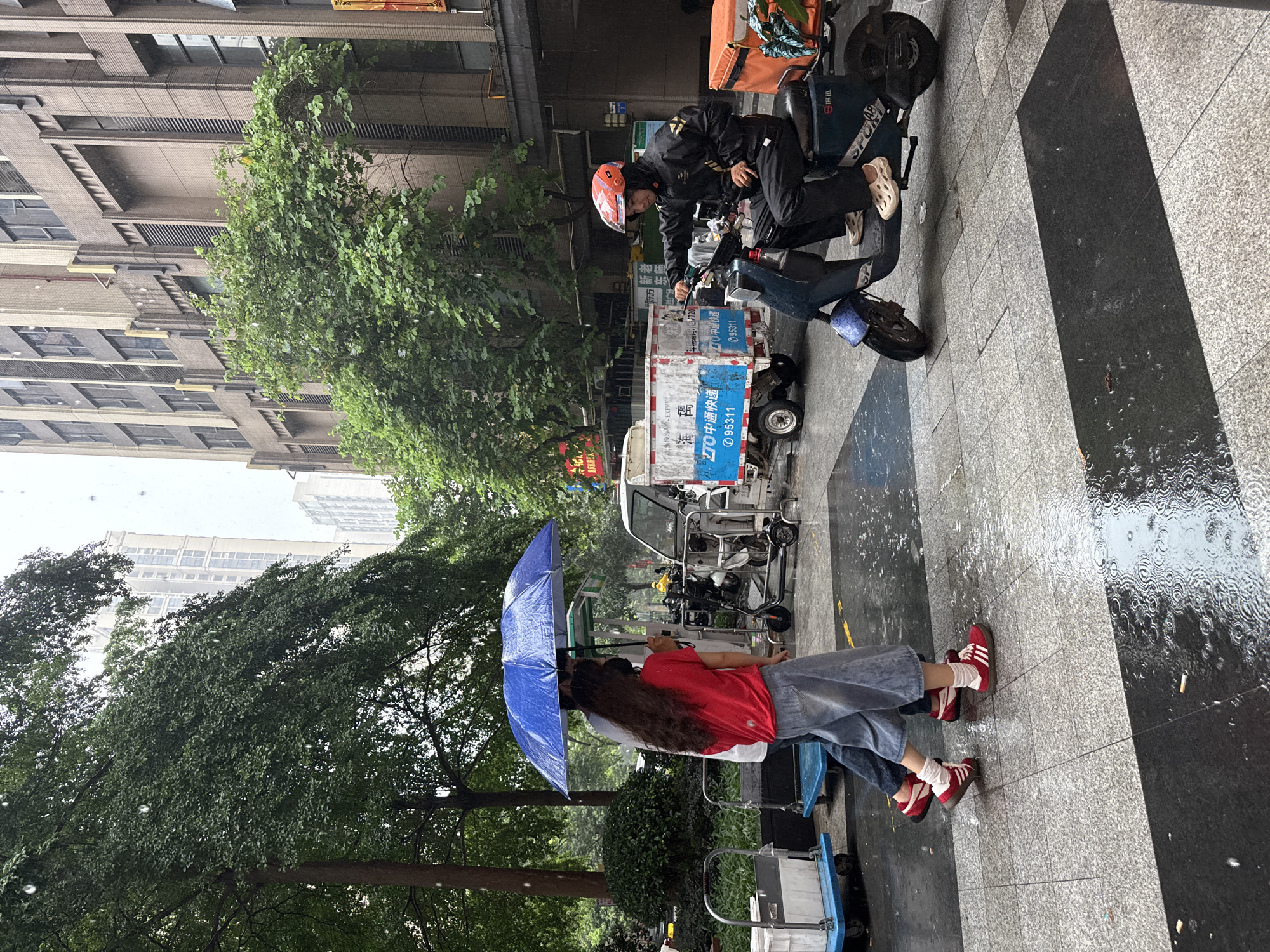 In Haizhu District, China, on a wet, overcast day, two individuals walk under a large blue umbrella while a delivery driver on a blue scooter waits at a red traffic light. The pavement is wet from the rain, with puddles reflecting the surroundings. In the background, a white delivery truck with Chinese characters, a building with a beige facade and vertical window elements, and lush green trees are visible. The person on the scooter wears a black hooded jacket and an orange helmet. The two pedestrians are dressed in casual clothing, with one wearing a red shirt and denim overalls, and the other in a white t-shirt and jeans. They are walking on a tiled sidewalk, with carts visible to the left of the frame.