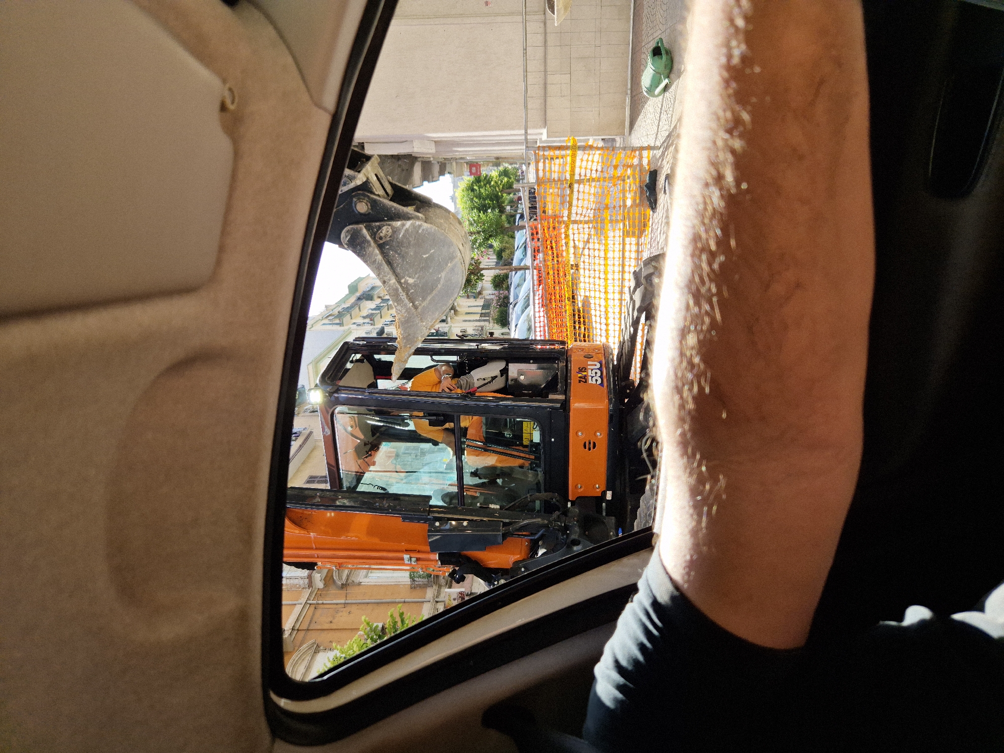 The image shows a view from inside a car, looking out the passenger window at a construction site. An orange excavator is visible, with a person in a yellow shirt operating it. The excavator is digging, and there is an orange safety fence around the construction area. In the background, there are buildings and parked cars. A green watering can is on the ground next to the fence. The scene appears to be on a street in Taranto, Italy.