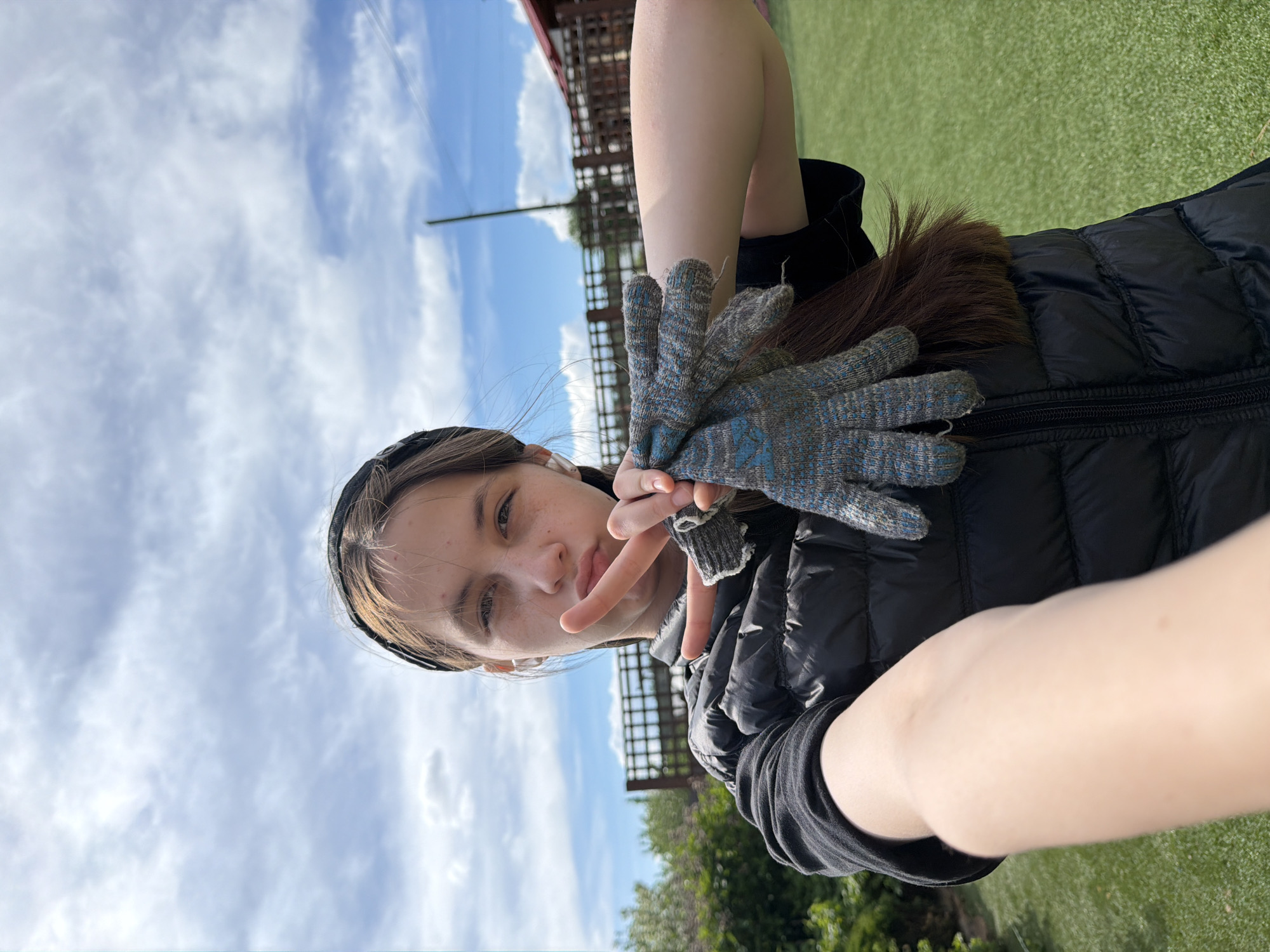 The image, originally rotated 90 degrees clockwise, captures a young person taking a selfie outdoors in Chaikovsky, Russia, during bright daylight.
The main subject is a fair-skinned individual with light brown hair, partially covered by a dark headband. They are wearing a black puffer vest over a dark long-sleeved top. Their left hand is raised, holding a pair of grey knitted gloves with blue patterns. Their right hand is also raised, forming a peace sign (V-sign) with two fingers extended. The person looks directly at the camera with a neutral expression, showing a slight pout and some visible freckles on their face.
The setting is outdoors with a clear blue sky dominating the upper part of the background, featuring scattered wispy white clouds. The lower part of the background shows green ground, which appears to be either natural grass or artificial turf. In the mid-ground, behind the person, faint dark structures, possibly part of a fence or a building, are visible, along with some distant foliage. The weather appears sunny and clear. There is no visible text in the image.