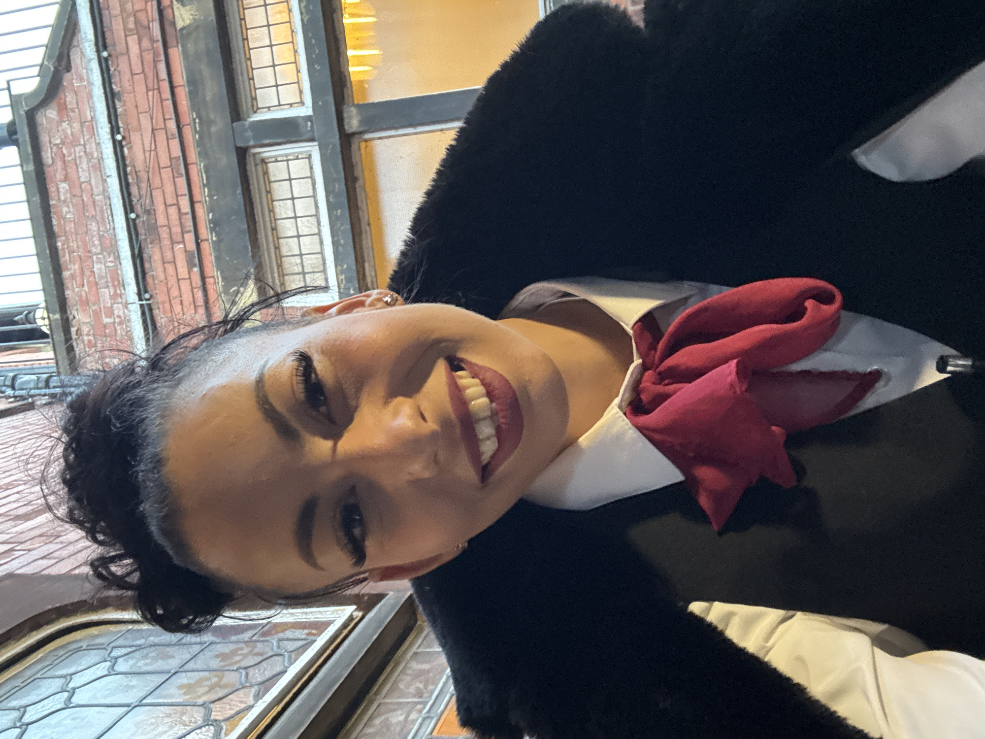 A woman with dark hair, fair skin, and wearing dark lipstick is smiling into the camera. She is wearing a white collared shirt, a dark coat, and a red neck scarf or bow. The background suggests an urban environment with a building constructed of red brick and white-framed windows. Based on the architectural style and the information provided, the photograph may have been taken in Warrington, United Kingdom.