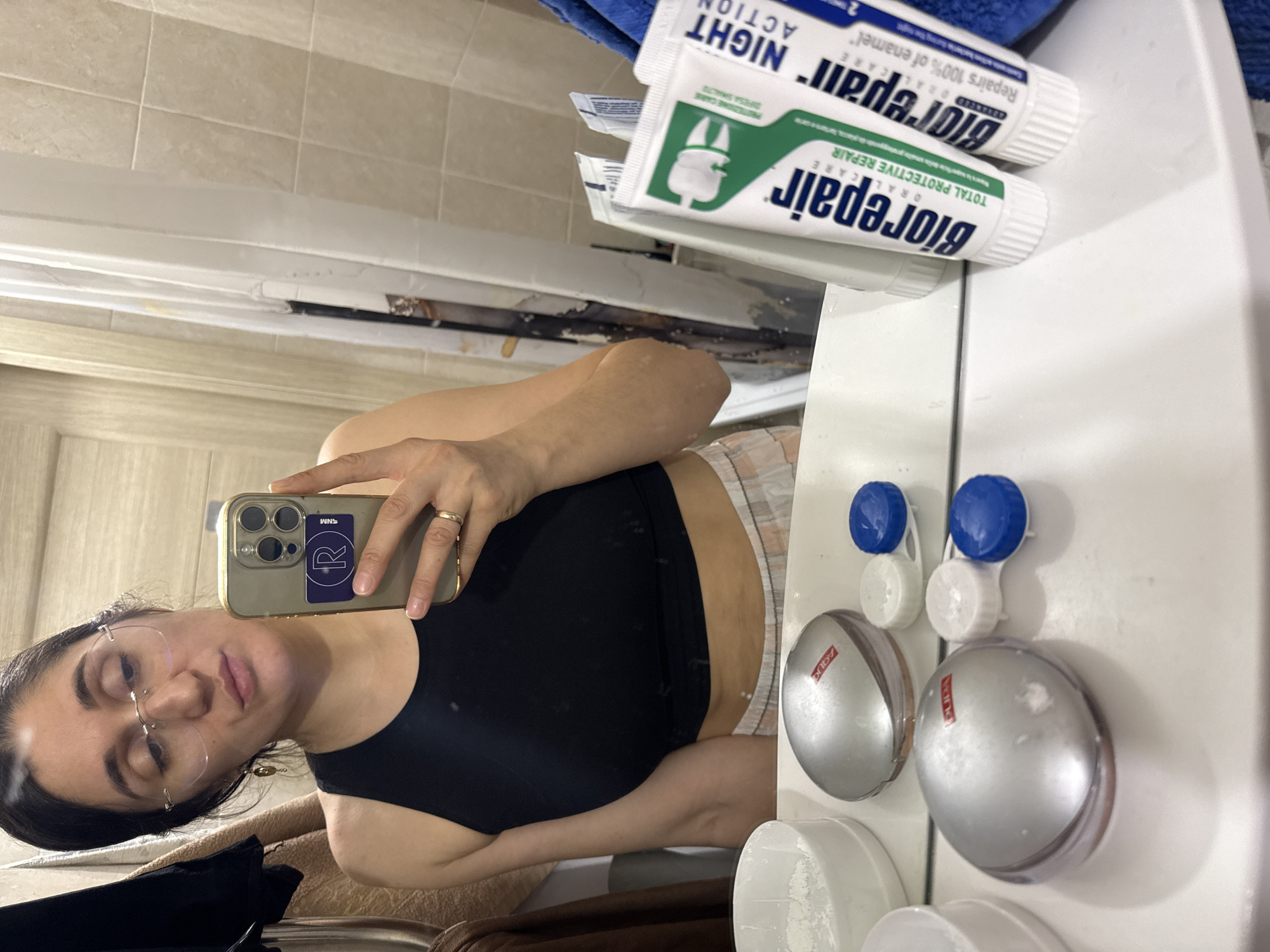 A young woman is captured in a mirror selfie within what appears to be a bathroom in Tobolsk, Russia. She holds a smartphone, featuring multiple cameras and a purple sticker with a white 'R' and "SNM" text, reflecting her image. She has dark hair, is wearing rimless glasses, a dark tank top, and light-colored plaid shorts. A ring adorns her left ring finger. Her expression is relaxed, with eyes partially closed, giving a casual or slightly weary impression.
The setting is a bathroom, identifiable by the tiled wall in the background and the white counter where various toiletries are placed. Prominently displayed are two tubes of Biorepair toothpaste: one labeled "TOTAL PROTECTIVE REPAIR" and the other "NIGHT ACTION," with visible text such as "Repairs 100% of enamel," "ORAL CARE," "PROTEZIONE CARIE," and "DIFESA SMALTO." Also on the counter are two contact lens cases with distinctive blue and white caps, and two round, silver compacts, one clearly marked "PUPA" and another with "AQUA." A blue towel is draped above the counter, and a brown textile is visible at the bottom of the frame, contributing to the personal, indoor ambiance.