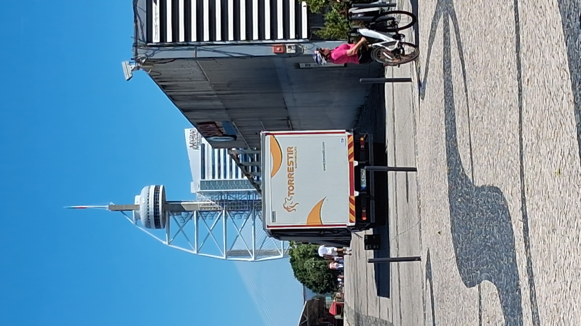 The image captures a sunny day in Lisbon, Portugal, near the Vasco da Gama Bridge and the Myriad Hotel. In the foreground, a person wearing a pink shirt and helmet rides a bicycle on a cobblestone path. To the left, a delivery truck with "TORRESTIR" branding is parked, its rear doors open. In the background, the distinctive white lattice structure of the Vasco da Gama Tower, a prominent landmark in the Parque das Nações area, stands tall against a clear blue sky. Several other buildings, including the Myriad Hotel, are visible behind it. A few people are walking in the distance, and shadows of objects and people are cast across the ground.