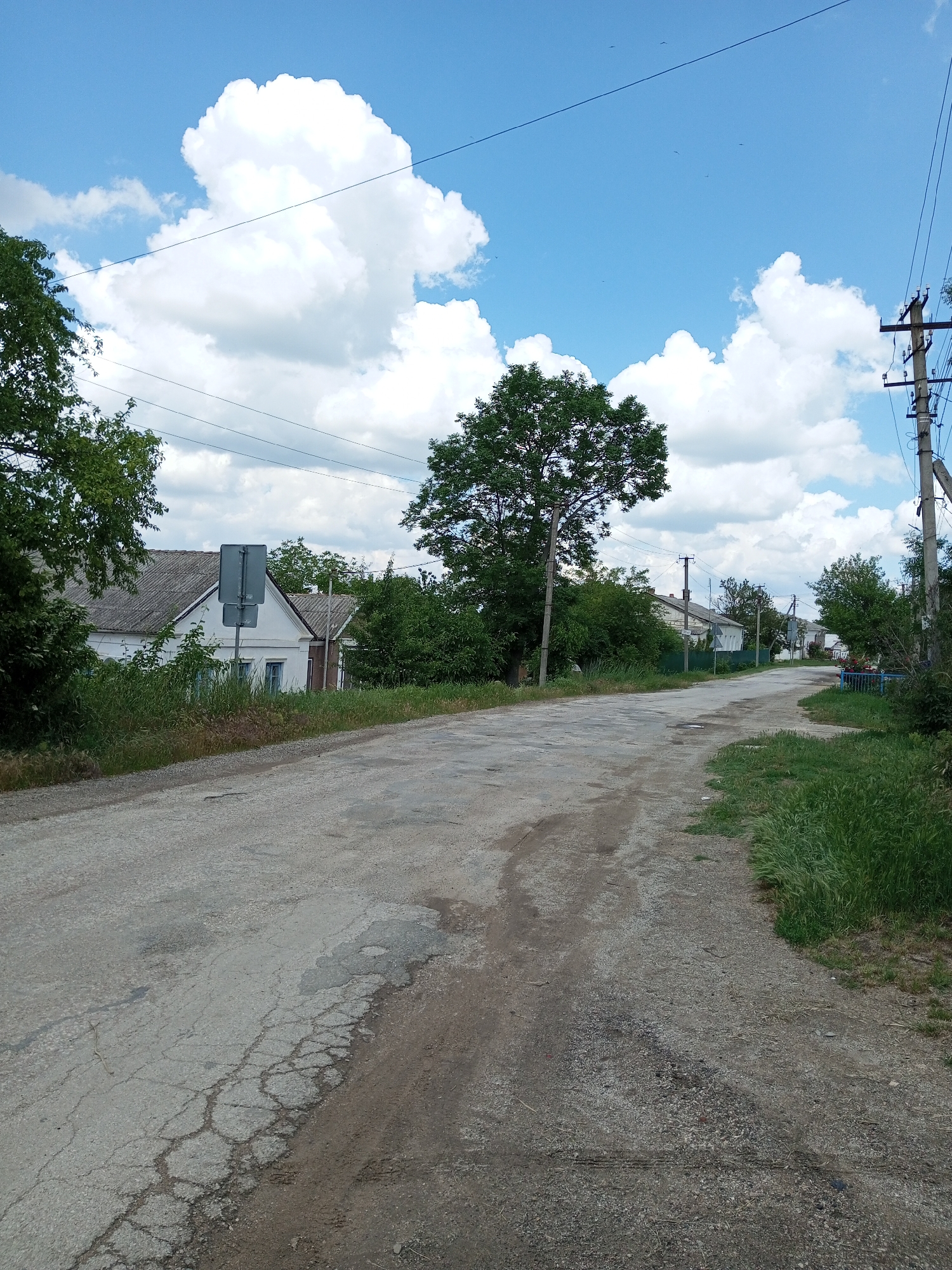 The image depicts a rural road in the Ароматновское сельское поселение, Ukraine. The road is made of asphalt and gravel and appears to be in disrepair, with visible cracks and patches. On the left side of the road, there is a white house with a gray roof, surrounded by overgrown vegetation and trees. A traffic sign is visible in front of the house. On the right side of the road, there are more trees, a power pole with electrical wires, and more houses in the distance. The sky is blue with scattered white clouds. The overall impression is of a quiet, somewhat neglected rural area. There are no people or animals visible in the image.