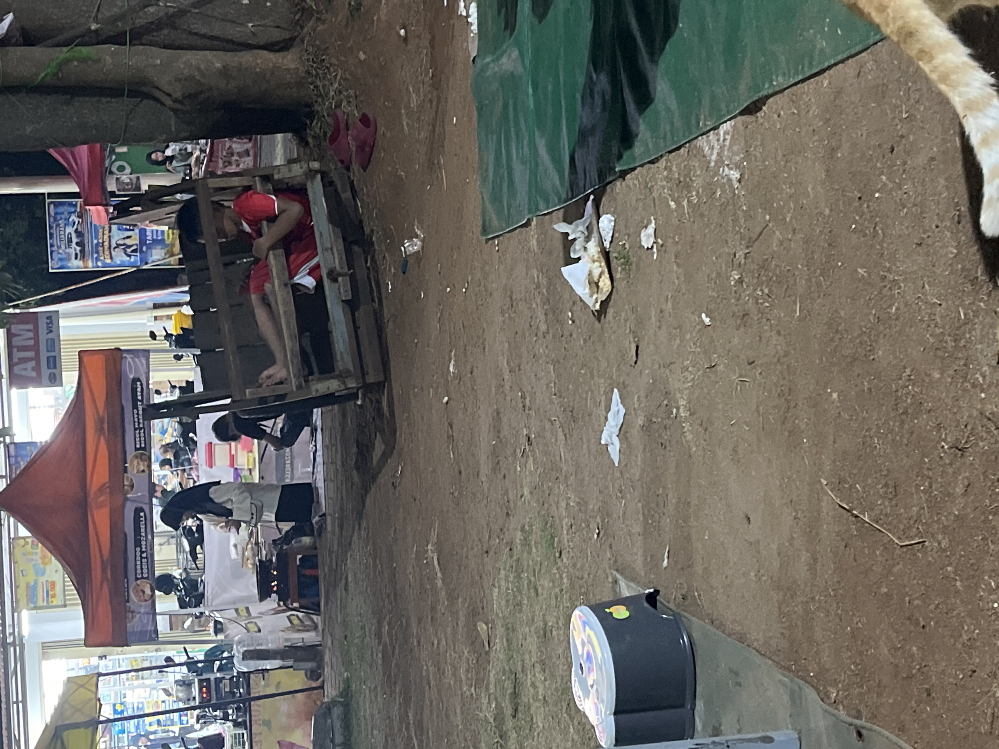 The image depicts an outdoor scene at night, possibly a food stall area, located in Cilegon, Indonesia. A young boy wearing a red t-shirt and shorts is seated on a wooden swing or bench, with his legs extended. To the left, a woman in a hijab and dark clothing is standing and appearing to cook at a small stove set up under an orange tent. Behind her, other stalls and a building with signage are visible. In the foreground, a green tarp is spread on the ground, and various debris, including food wrappers and plastic, are scattered around the area. A small, dark plastic stool with colorful decals is also present. The ground is covered with dirt and patches of grass. The overall impression is a casual, possibly informal, nighttime market or gathering space.