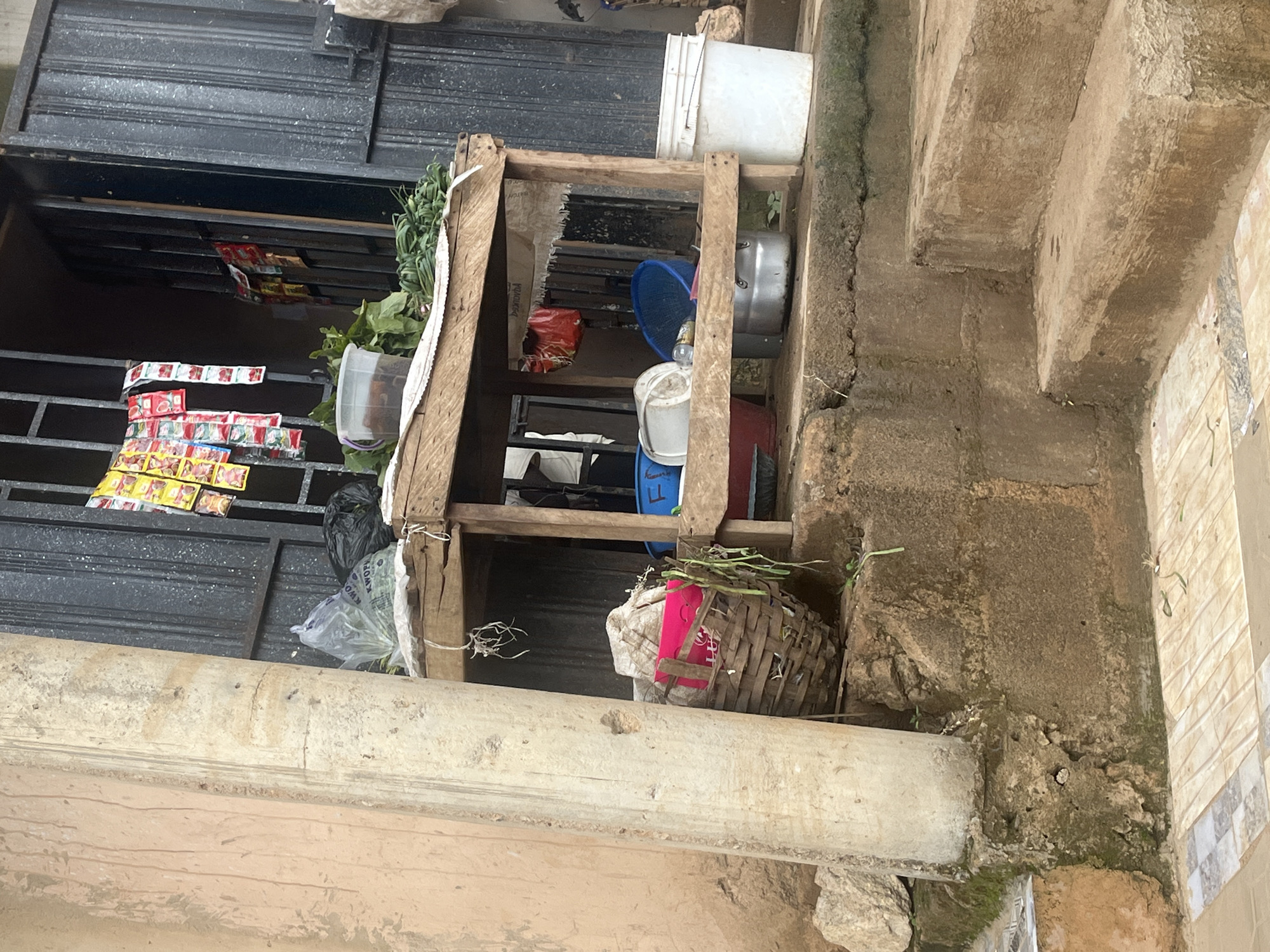 The image depicts an outdoor scene in Gura Top, Nigeria, likely a marketplace or street vendor stall. In the foreground, a rough concrete staircase leads up to a wooden stall. To the left, a large, roughly textured pillar stands next to the stairs. The stall itself is cluttered with various items. On a shelf, there are bunches of green vegetables, a clear plastic container with liquid, and a black plastic bag. Below the shelf, a woven basket contains items, including a bright pink object. Further to the right, a white bucket and a metal pot are visible. In the background, a dark, barred doorway or entrance suggests a building. Small packets of colorful items are hanging from the bars above the stall, possibly indicating goods for sale. The overall impression is one of a functional, everyday commercial space in an urban setting.
