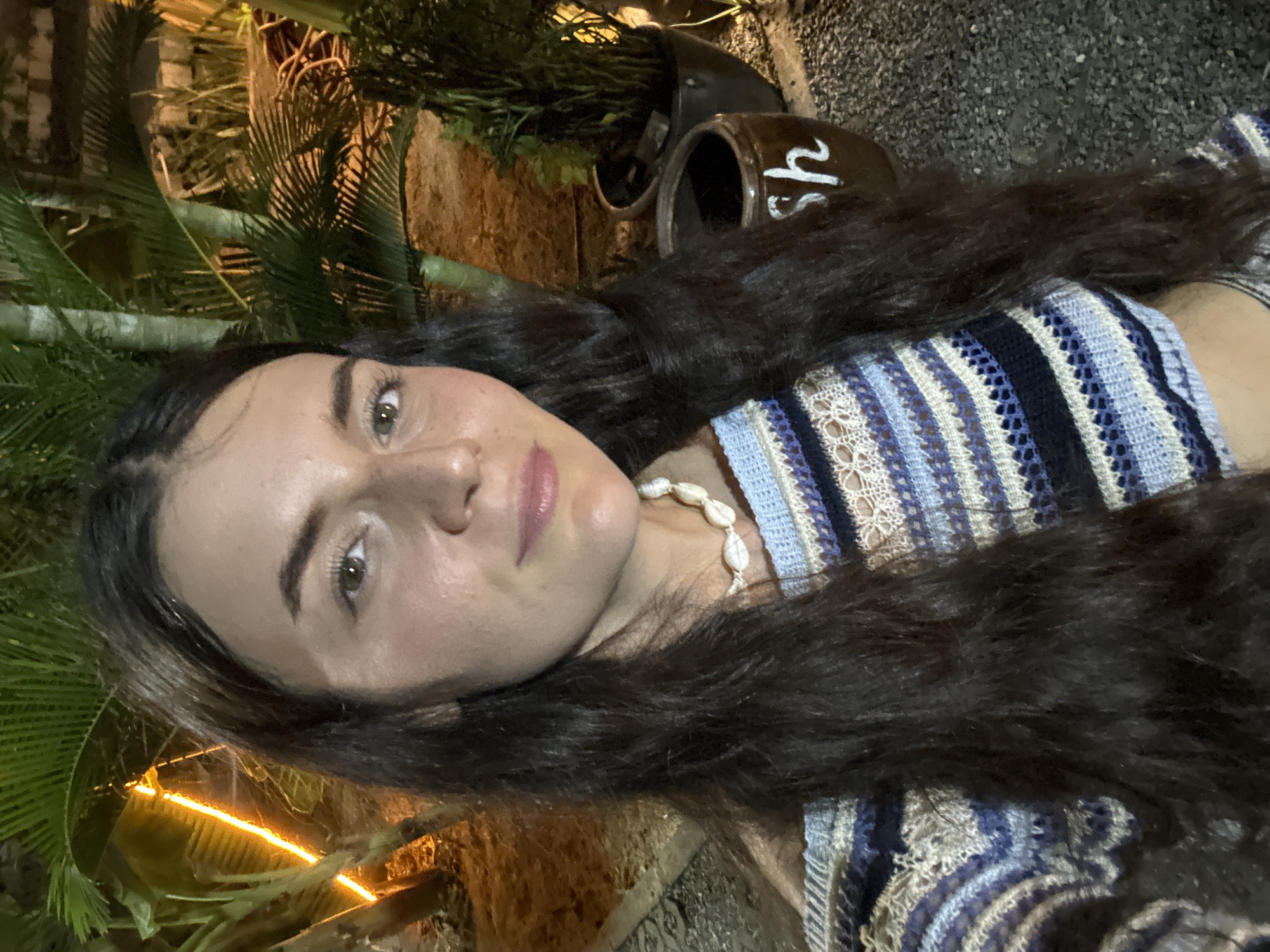 A woman with long, dark hair lies in an outdoor setting. She wears a striped blue and white crochet top and a seashell necklace. She is positioned among tropical plants, including what appears to be palm trees. An earthenware pot with white lettering sits near her. The environment has an outdoor, possibly evening, ambiance. The specific location in Mũi Né, Vietnam, is not identifiable from the image alone.