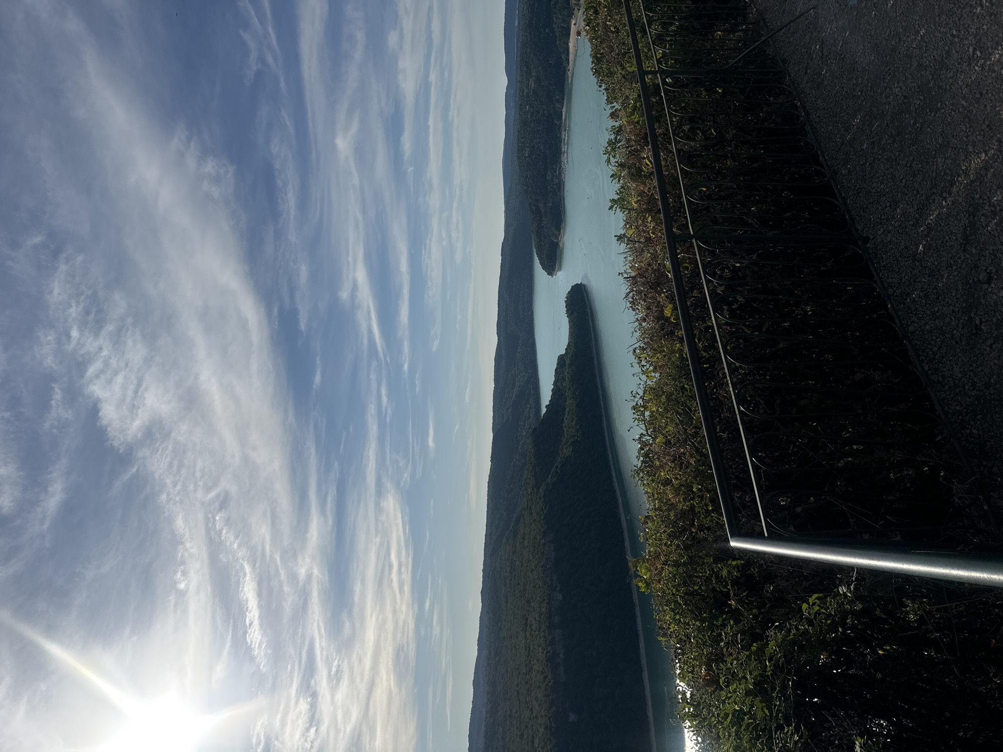 An aerial shot captures a scenic view of a wide, winding body of water, likely a lake or reservoir, surrounded by lush, tree-covered hills. The sun shines brightly in the upper left quadrant of the frame, casting a lens flare and illuminating the wispy clouds that streak across a clear blue sky. In the foreground, a dark metal railing borders a dense green bush line, which overlooks the expansive waterway. The water itself is a tranquil turquoise, reflecting the daylight and the surrounding foliage. Boats can be spotted dotting the surface of the water, and the terrain on the opposite shore appears more populated with buildings visible amidst the trees. The overall impression is one of natural beauty and serene leisure, potentially a viewpoint above the Lac de Vouglans near Moirans-en-Montagne, France, a popular tourist destination known for its vast reservoir and surrounding natural landscapes.