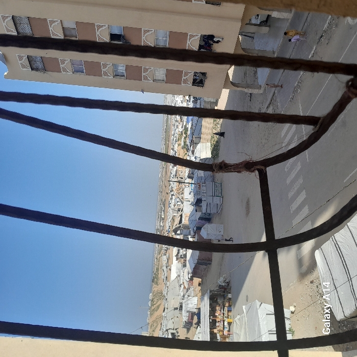View from a high vantage point, possibly a window behind a decorative iron grill. A street with pedestrian crossings runs below, alongside a dense encampment of tents and makeshift dwellings stretching into the distance. Adjacent to the window, a tall building with multiple floors and windows is visible. The sky is clear and light blue. A "Galaxy A14" watermark is apparent at the bottom right corner.