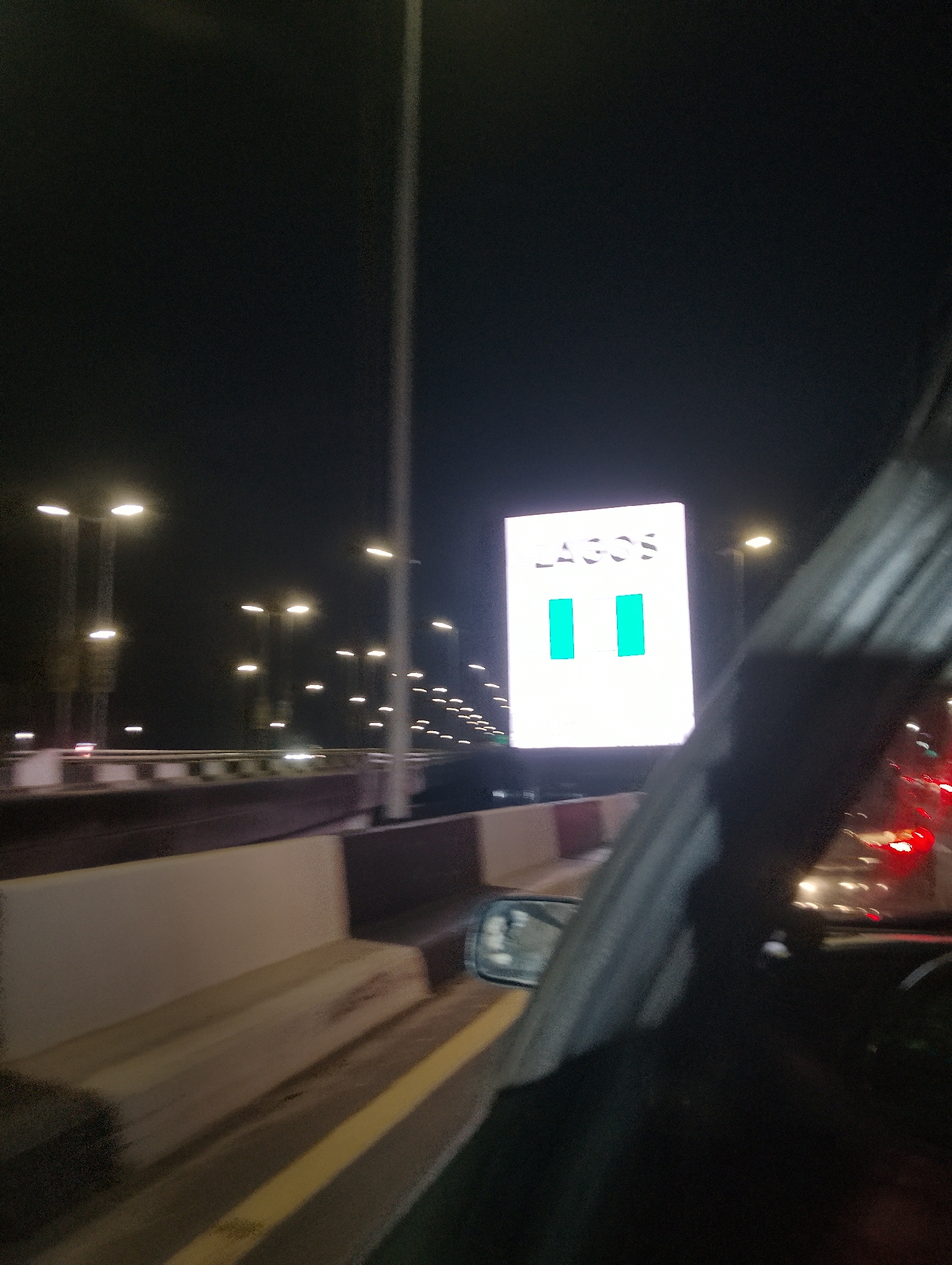 An aerial view shows the busy streets of Ikoyi, Nigeria, at night. The city is illuminated by numerous streetlights and a large, brightly lit digital billboard displaying the word "LAGOS" in large white letters, with two teal-colored rectangles below. The billboard is positioned above a highway, with traffic visible in the distance. The image is captured from within a moving vehicle, indicated by the presence of a side mirror and the blurred motion of the car's interior. The highway has concrete barriers along its sides, and the overall scene suggests a vibrant urban environment.