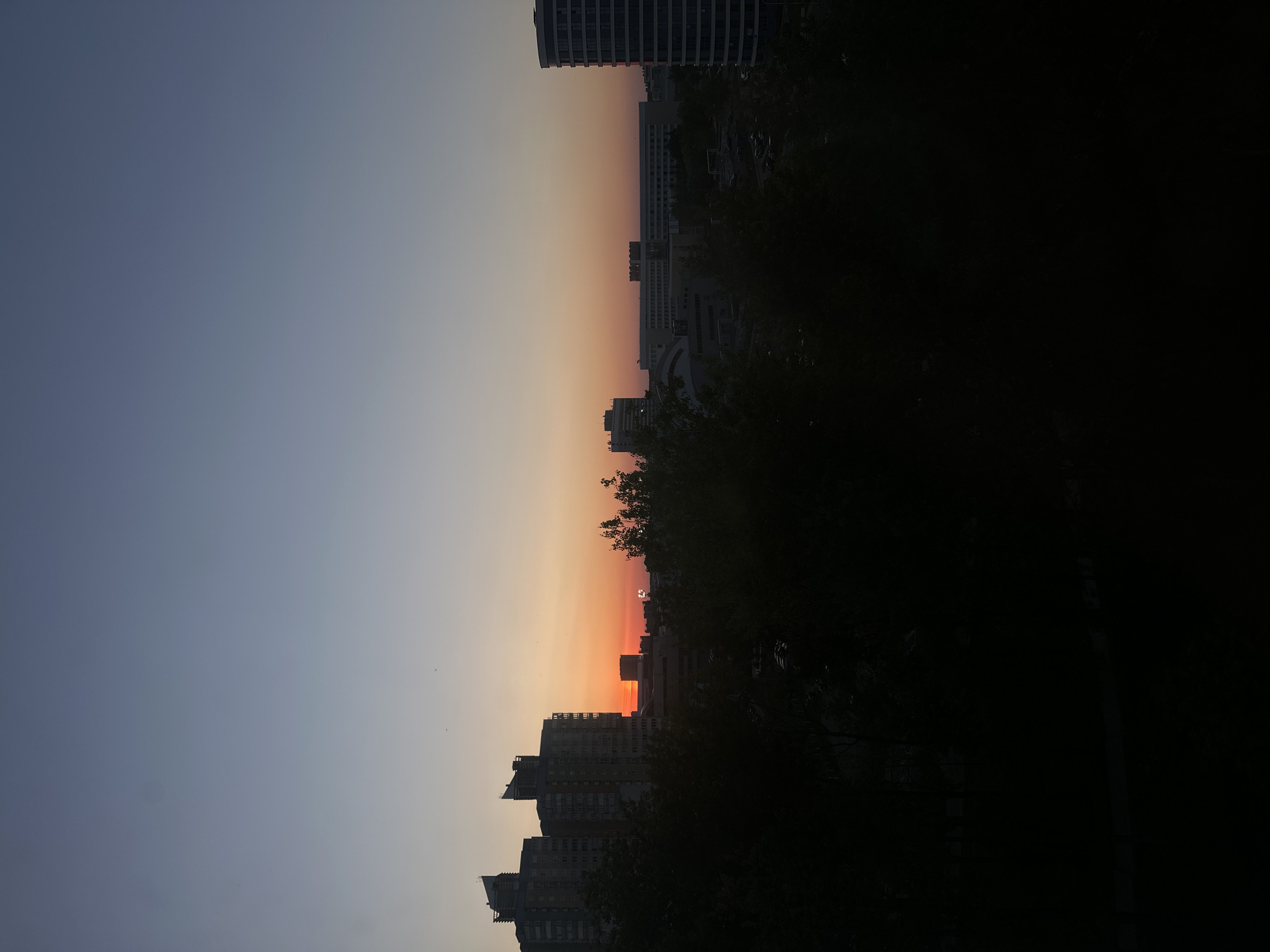 The image captures a sunset scene in Minsk, Belarus, featuring a cityscape. Buildings and trees are silhouetted against a gradient sky, ranging from dark blue to orange near the horizon. The darkness of the foreground suggests the presence of trees or structures obscuring a direct view. The perspective indicates an elevated vantage point, likely from a building or high ground. The image lacks human presence and focuses on the interplay of natural light and urban infrastructure.