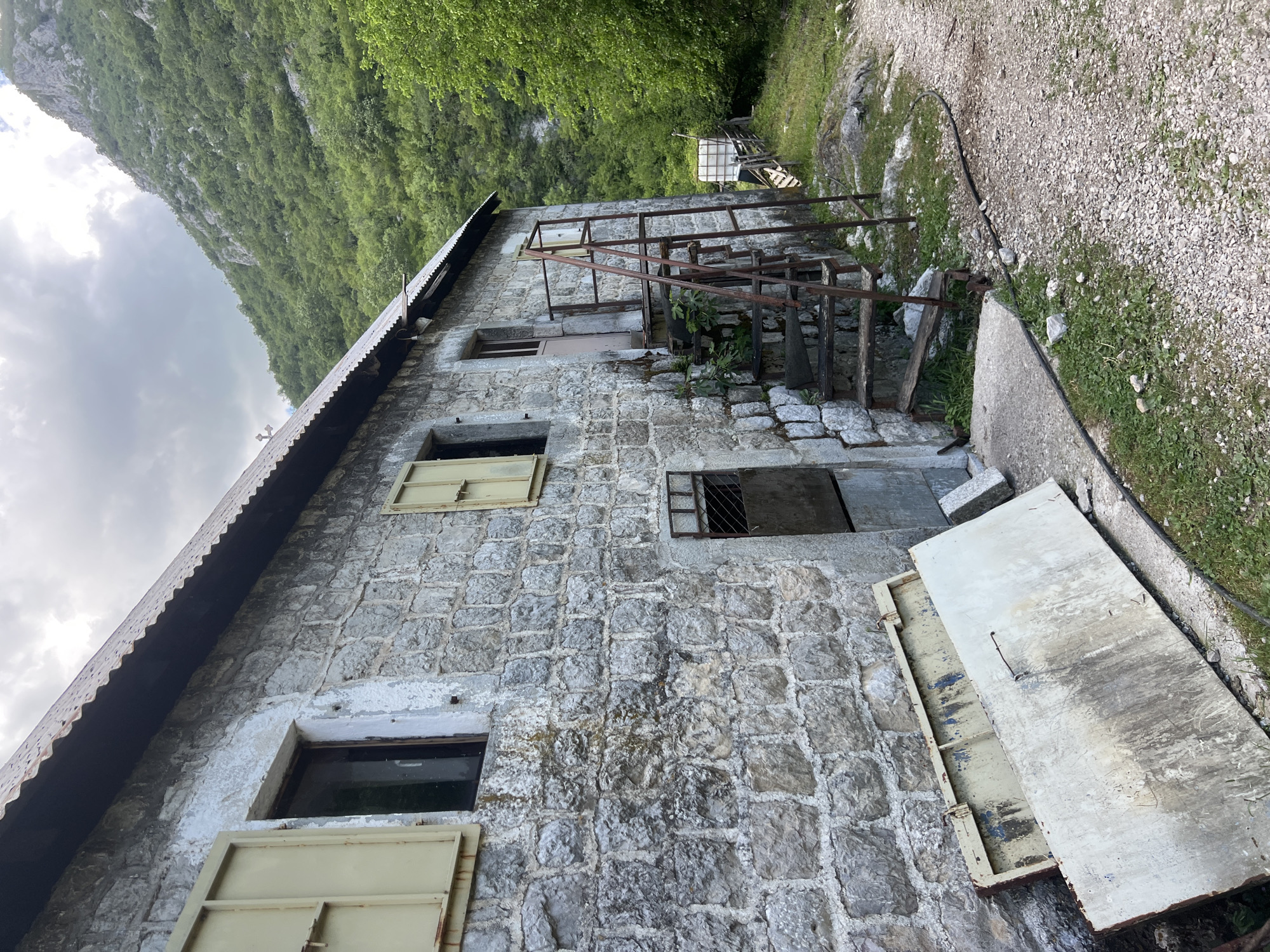 The image shows the exterior of a two-story stone building in Mrtvo Duboko, Montenegro. The building has a corrugated metal roof and several windows, some with metal shutters. A metal staircase leads to the second story. The surrounding environment includes a gravel path, vegetation, and a mountain in the background. A large white metal door is lying open on the ground next to the building. There are no people visible in the image.