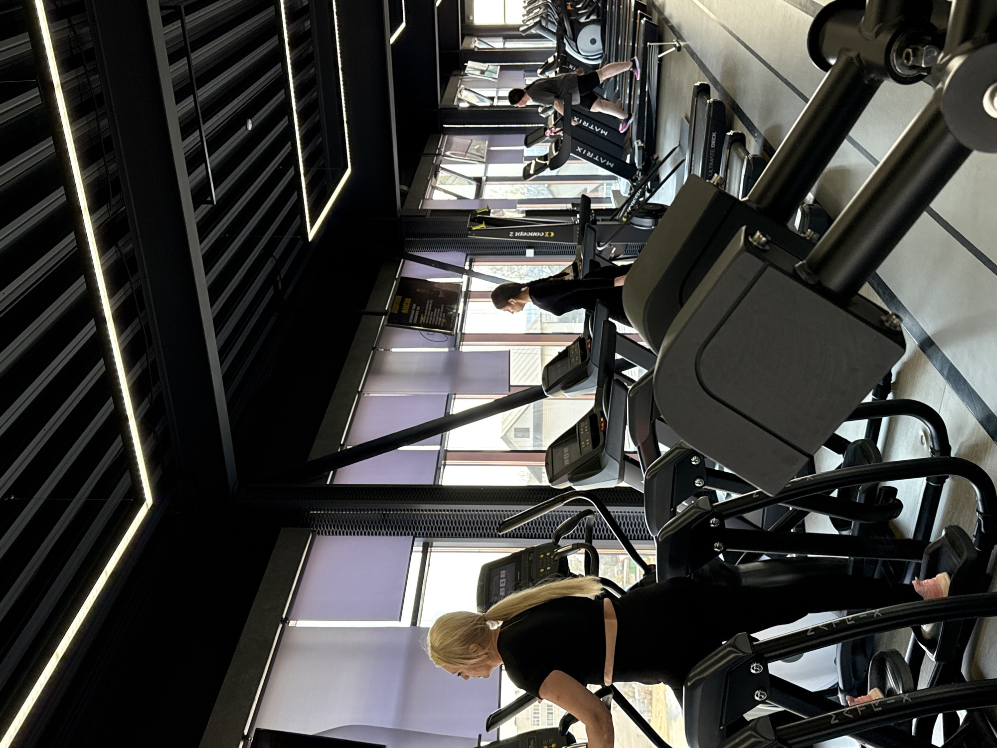 The image displays a modern gym interior where three individuals are engaged in cardiovascular exercise. In the immediate foreground, a woman with blonde hair tied in a ponytail, wearing a black t-shirt and leggings, is intently focused on an elliptical machine. This machine, and others nearby, feature the branding "2FLEX".
Further into the gym, another person with short dark hair, dressed in a dark top, is also using a piece of cardio equipment, possibly a rowing machine, with "CONCEPT2" visible on adjacent gear. In the background, a man wearing a grey t-shirt and shorts, noticeable for his pink-soled shoes, is exercising on a treadmill. The treadmills are clearly branded "MATRIX".
The gym's design is contemporary, featuring high ceilings with dark, exposed structural beams and prominent linear LED light strips that illuminate the space. Large windows line one wall, allowing natural light to enter and offering a glimpse of an outdoor scene with buildings under an overcast or bright sky, suggesting it is daytime. The windows are partially obscured by light-colored blinds. The walls are light-toned, possibly with subtle horizontal stripes.
The overall atmosphere is one of focused individual activity. The equipment is predominantly dark, contrasting with the bright lighting and the light tones of the walls and natural light. A screen or poster in the mid-ground is too blurry to decipher most text, though "EDITION" is faintly visible.
