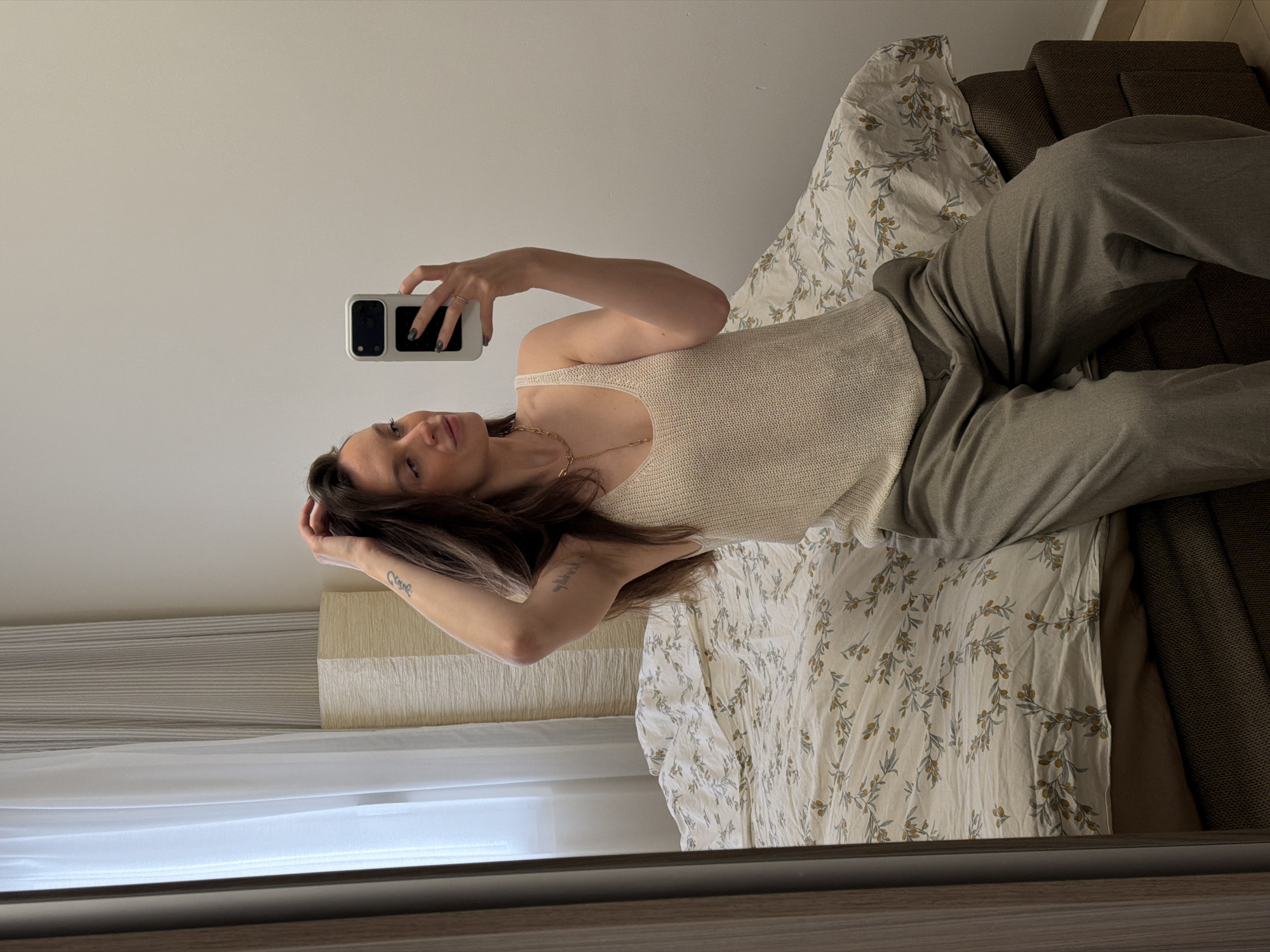 A woman is taking a selfie in a mirror. She is lying on a bed with a floral pattern duvet cover. The bed is next to a striped wall and a dark brown headboard. She is wearing a beige tank top, trousers, and a gold necklace. She has dark hair and a tattoo on her left forearm. She is holding a white smartphone. The background suggests the location is an interior space with natural light, but there are no indicators to identify this specifically in Budva, Montenegro.