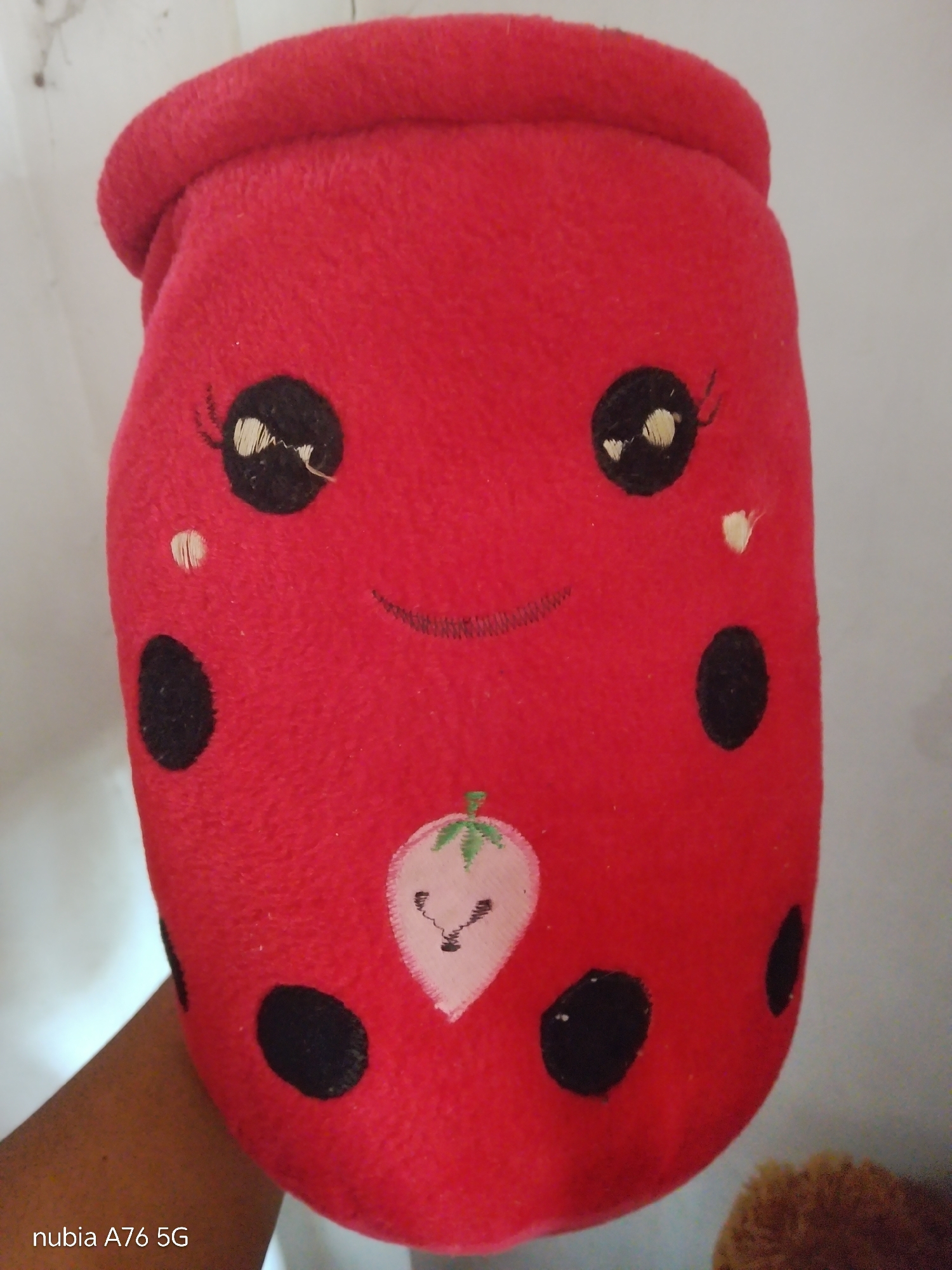 A close-up, eye-level shot shows a plush toy designed to resemble a red ladybug. The toy is elongated and appears to be cylindrical, with a rounded top that has a rolled hem. It has two large, black, circular eyes with white highlights and eyelashes. A small, stitched smile is positioned below the eyes. On the front of the toy, a light pink strawberry with a green stem and leaves is embroidered. Black spots, characteristic of a ladybug, are scattered across the red surface. A hand is visible on the left side of the frame, holding the toy. The background is a plain, light-colored wall. The image is located in Cakung, Indonesia.