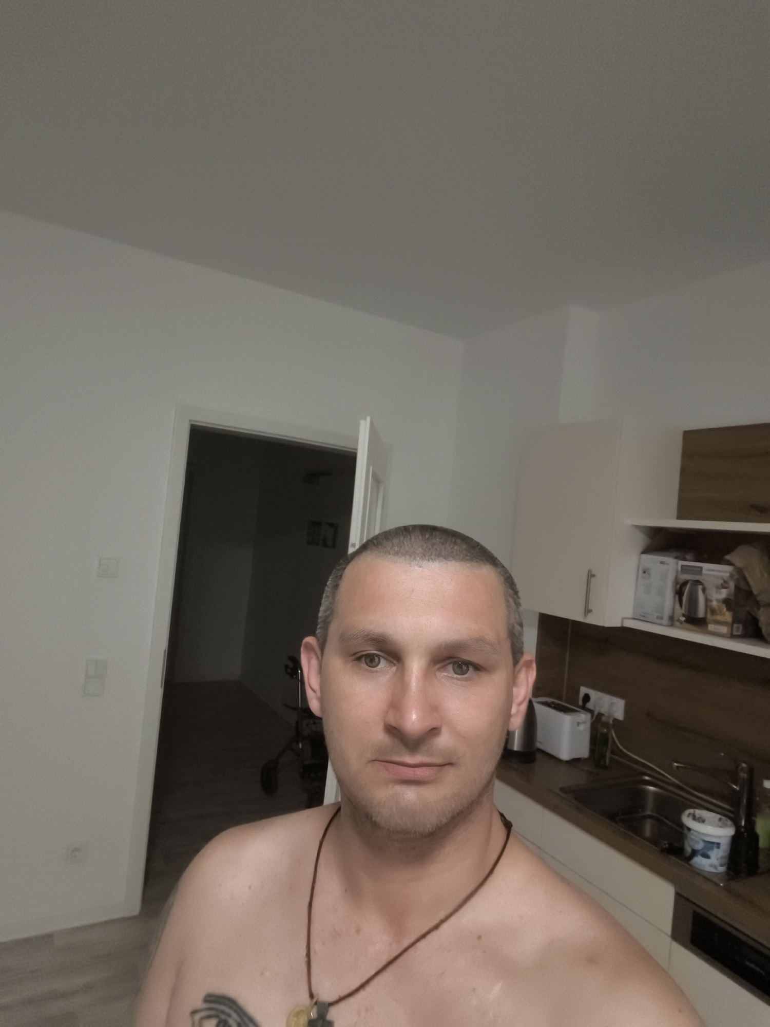 A man with short, graying hair and stubble is photographed from the chest up, indoors. He is wearing a necklace. To his left, a doorway reveals a hallway with a walker. To his right, a kitchen counter is visible with a sink, a toaster, a kettle, and various containers. The walls and cabinets are white. The scene appears to be a domestic interior, likely a kitchen and adjacent hallway.