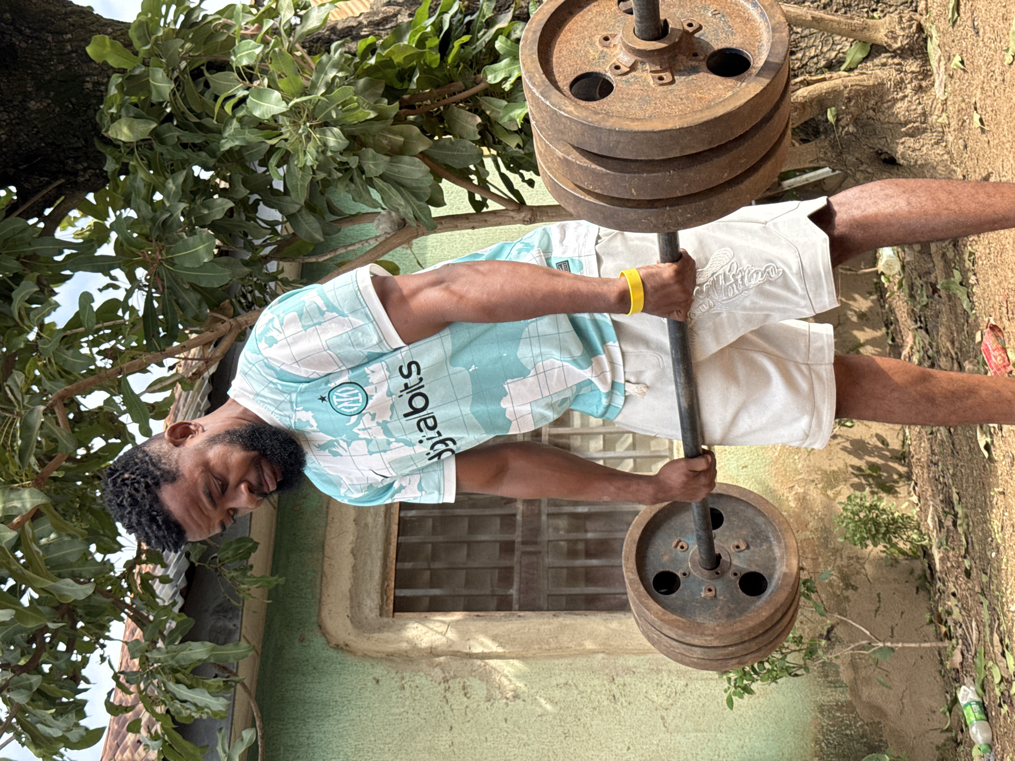 A Black man is performing a deadlift outdoors in Akwanga, Nigeria. He is wearing a light blue and white patterned t-shirt with "digibits" written on it, white shorts, and a yellow wristband. He is gripping a barbell loaded with several heavy, rusty weight plates. The background shows a simple, weathered building with barred windows, overgrown foliage, and a large tree. The ground is covered with dirt, leaves, and a discarded plastic bottle. The lighting suggests it is daytime.