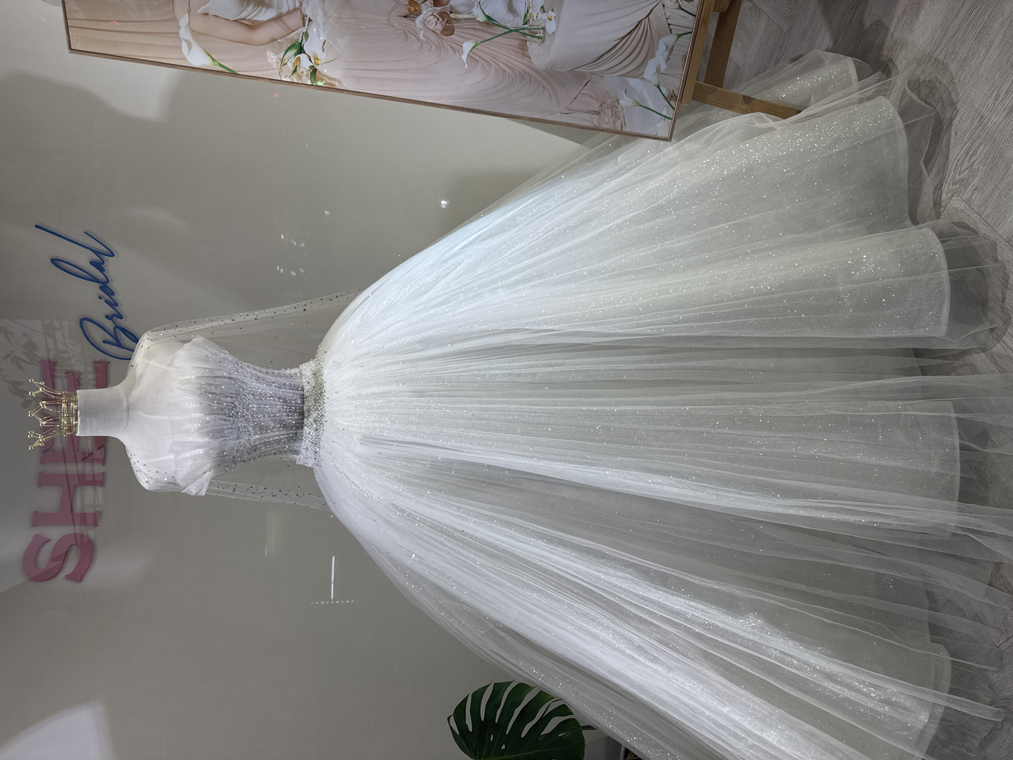 The image shows a white wedding dress displayed on a mannequin in what appears to be a bridal shop. A gold crown is placed atop the mannequin's head. The dress features a beaded or sequined bodice and a full, layered skirt of glitter-flecked tulle. The background includes a framed image of a bride and a display with the word "Bridal" written in a blue cursive font. Additionally, a pink sign is visible behind the mannequin. A green potted plant is placed at the bottom of the mannequin. The location appears to be Đà Nẵng, Vietnam, considering the She Bridal advertisement. The floor is covered with a light gray, possibly wood-effect, material.