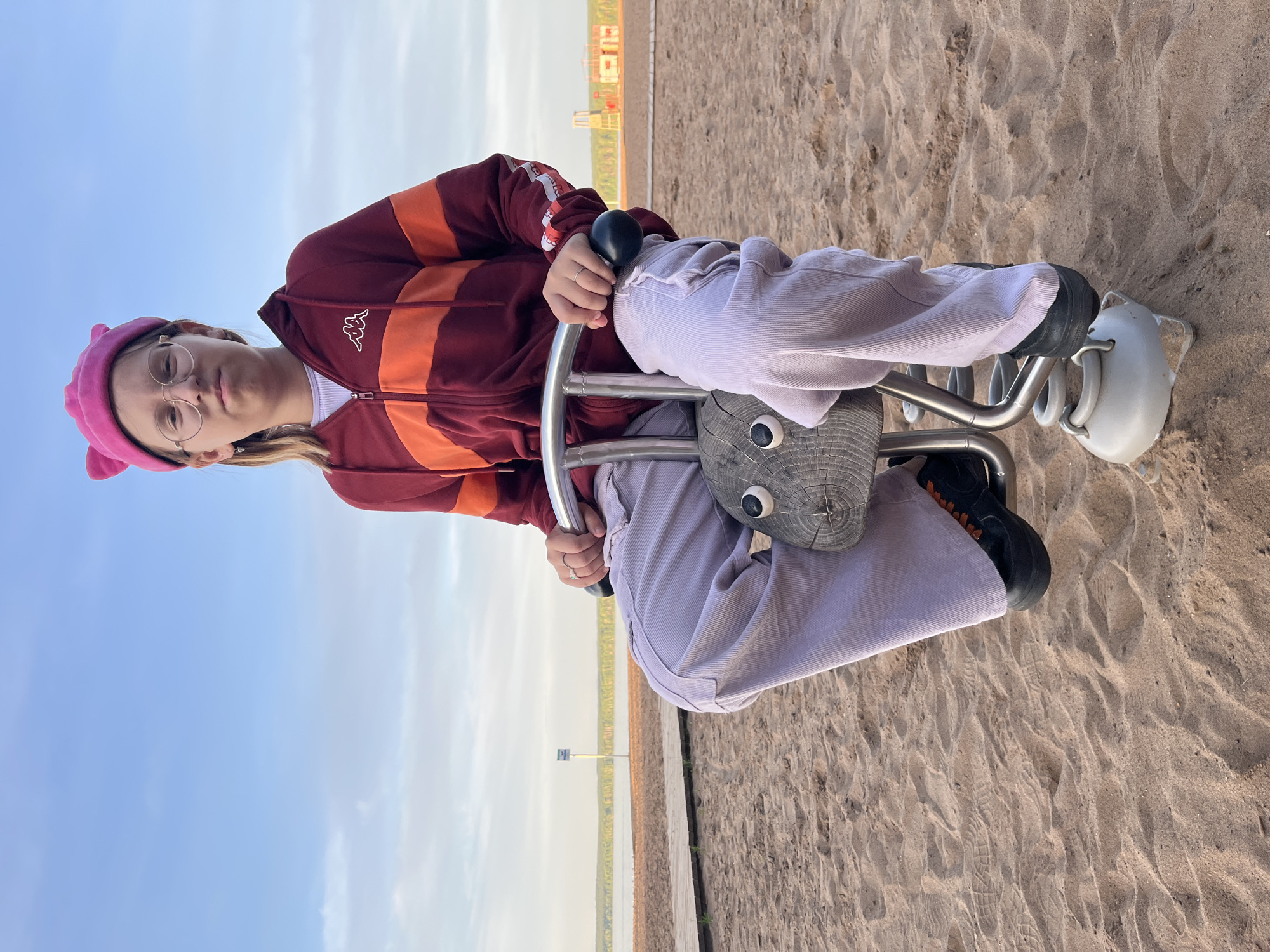 A young individual is pictured sitting on a spring rider playground toy. The person, positioned facing the camera, has a neutral expression and wears round eyeglasses, a pink beret-style hat (possibly with small ears), and a maroon and orange striped zip-up jacket that features a white "KAPPA" logo on the chest. They are also wearing light-colored corduroy pants and dark shoes. The spring rider is made of wood, shaped like a stylized animal head with prominent circular eyes, and is mounted on a metal spring base with black handles which the person is holding.
The setting is an outdoor recreational area. The foreground consists of sandy ground, showing footprints. Behind this sandy area, a wooden boardwalk or path stretches horizontally across the scene. Beyond the path, a large body of water, likely a river or lake (consistent with the Kama River in Chaikovsky, Russia), extends into the distance. The far shoreline features some indistinct buildings and a tall mast or pole. The sky is bright blue with a few scattered clouds, indicating clear, daytime weather. The individual appears to be casually enjoying the playground equipment.