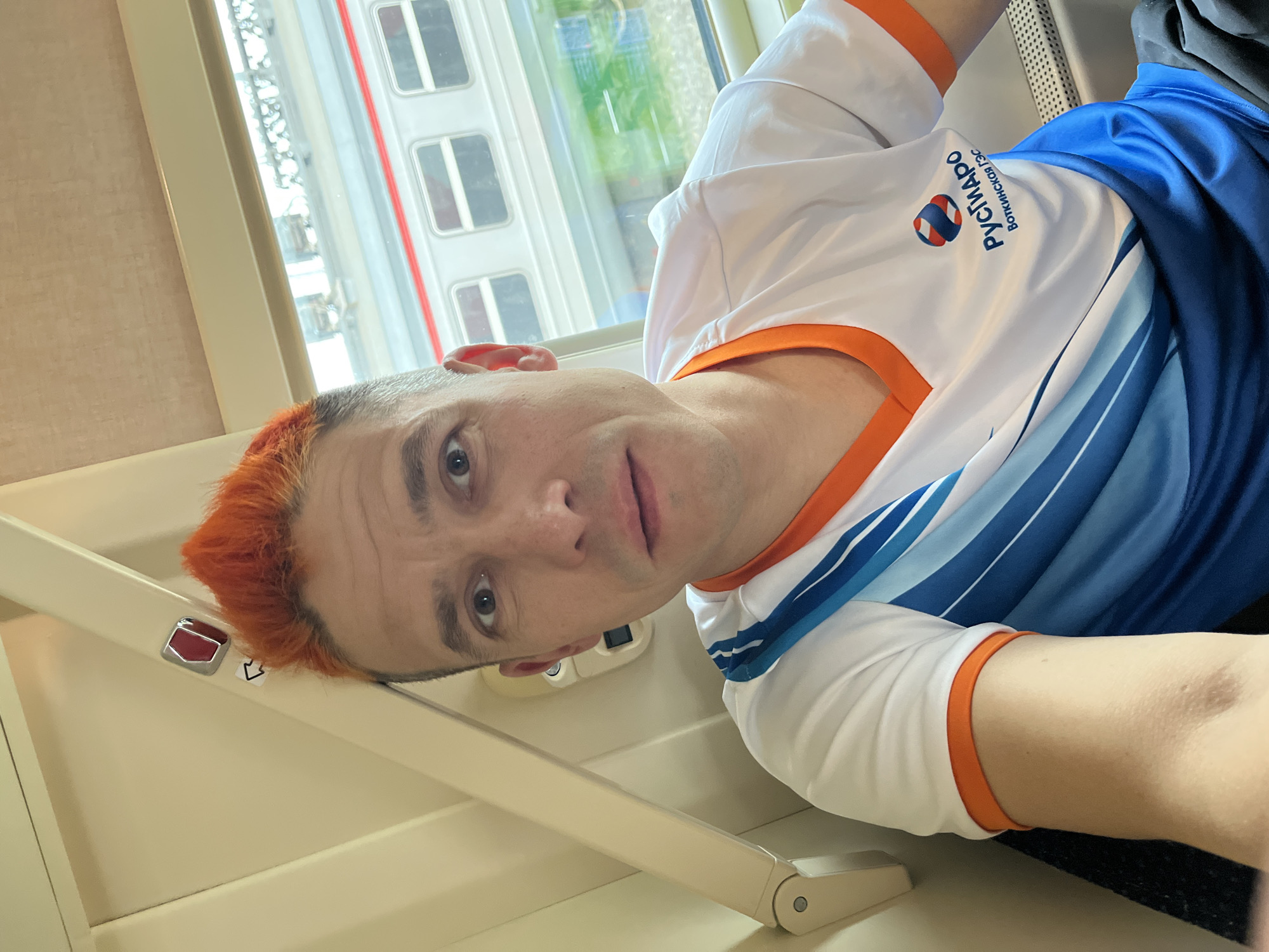 A man with red hair is inside a train, possibly on a journey within Izhevsk, Russia. He is wearing a white shirt with orange trim and a blue design, featuring a logo in Russian, "РУСГИДРО Воткинская ГЭС" (RusHydro Votkinskaya HPP). The train window shows a view of buildings outside. The man is looking at the camera. He may be traveling for reasons related to the RusHydro Votkinskaya HPP power plant.