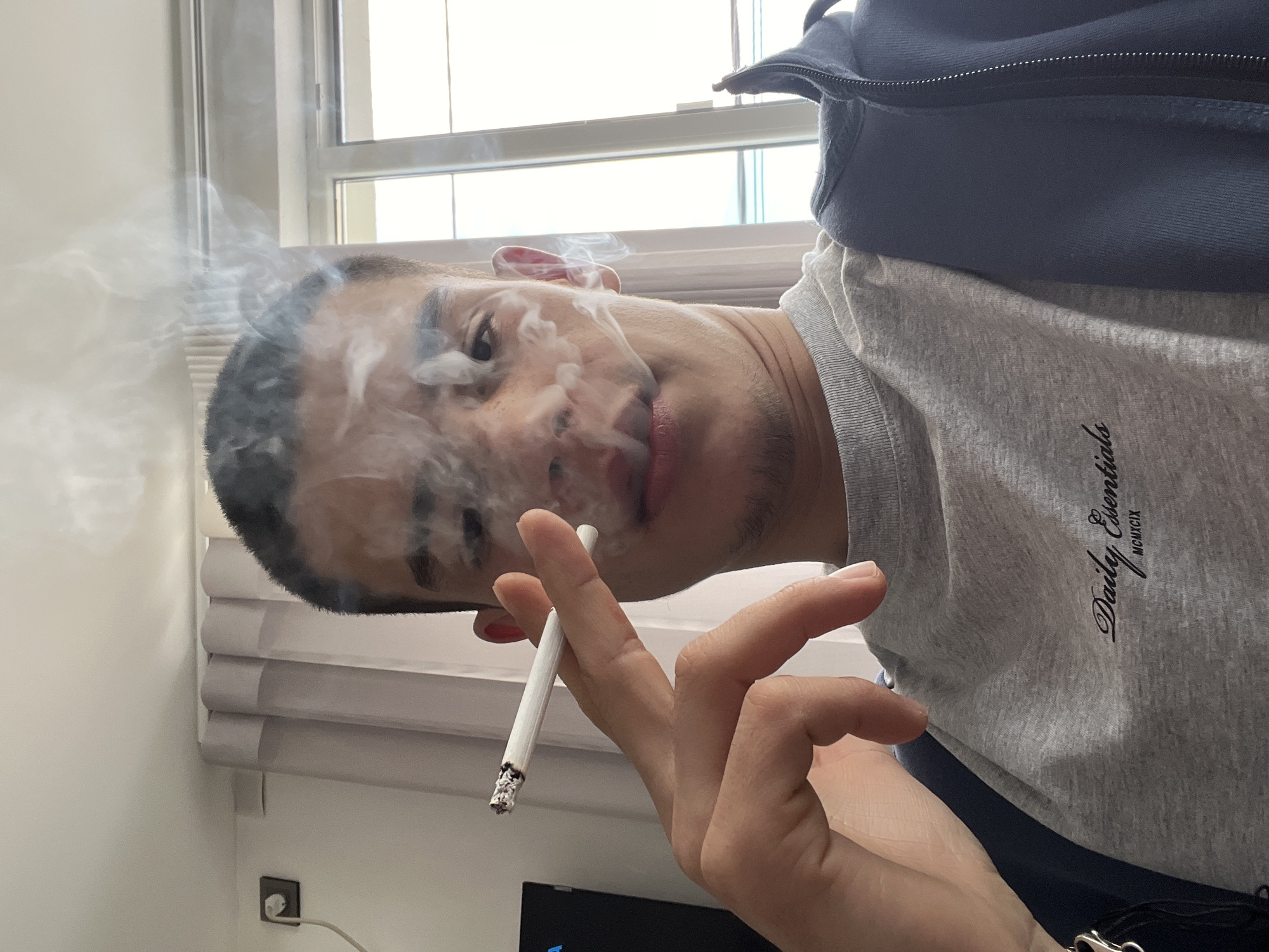 A man is depicted holding a lit cigarette in his right hand. Smoke is rising from the cigarette and partially obscures his face. He is wearing a grey t-shirt with the inscription "Daily Essentials MCMXCIX" on the right side of his chest and a navy jacket. The scene appears to be indoors, with a window and blinds visible in the background. An electrical outlet is visible on the wall behind the window. There is no evidence to suggest the location is Karavelovo, Bulgaria.