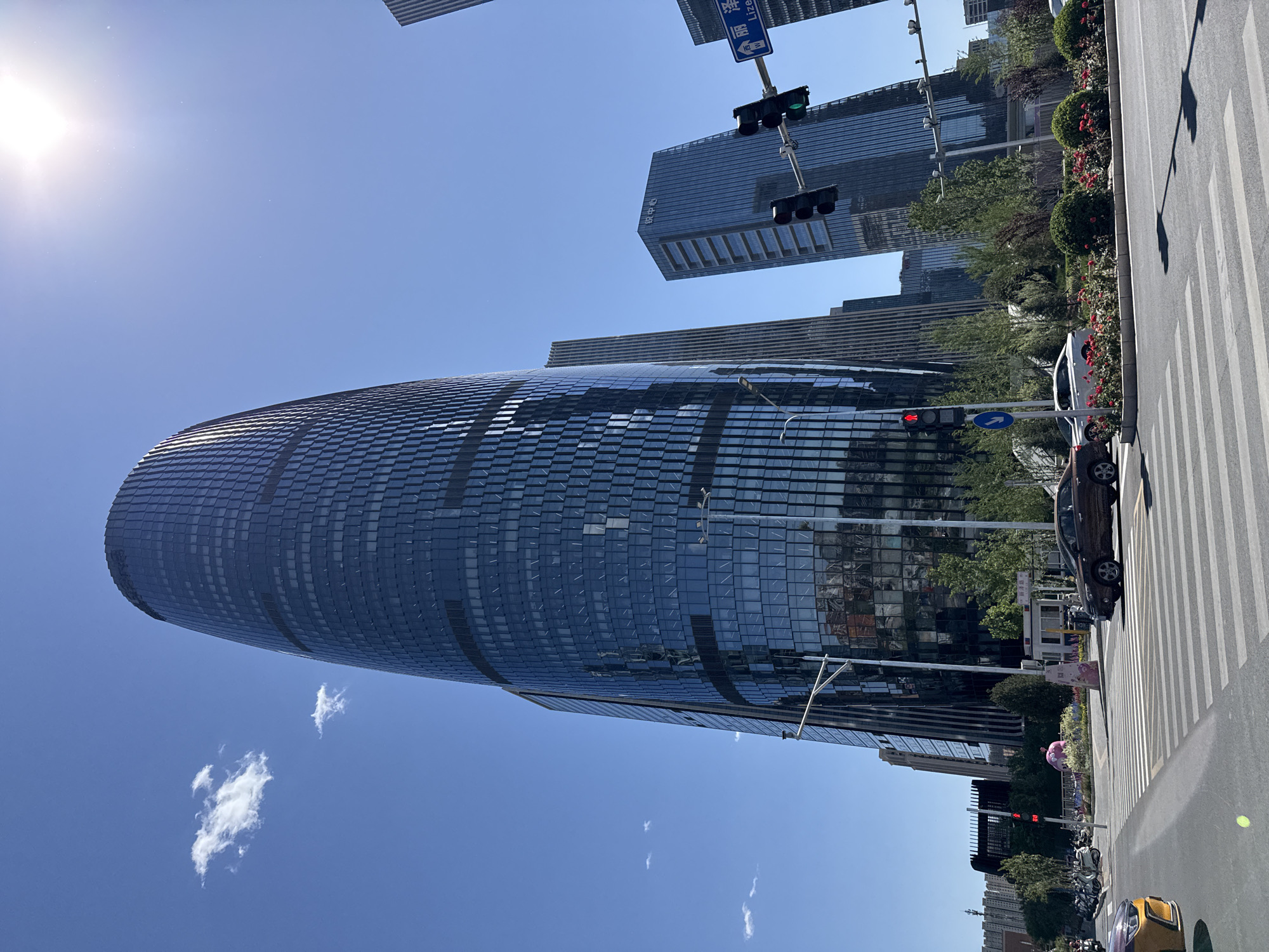 The image depicts a street scene in Fengtai District, China, featuring modern architecture and urban elements. A cylindrical skyscraper dominates the left side of the frame, its glass facade reflecting the clear blue sky with scattered clouds. To the right, a more conventional, rectangular skyscraper is visible. A street runs along the right edge of the image, complete with a pedestrian crosswalk, traffic lights (showing green and red), and a direction sign. Motor vehicles, including cars and motorbikes, are visible along the street, alongside a flowerbed or landscaping. A person holding a pink balloon is also seen on the sidewalk. The lighting suggests a sunny day, with the sun's glare visible in the upper-left corner.