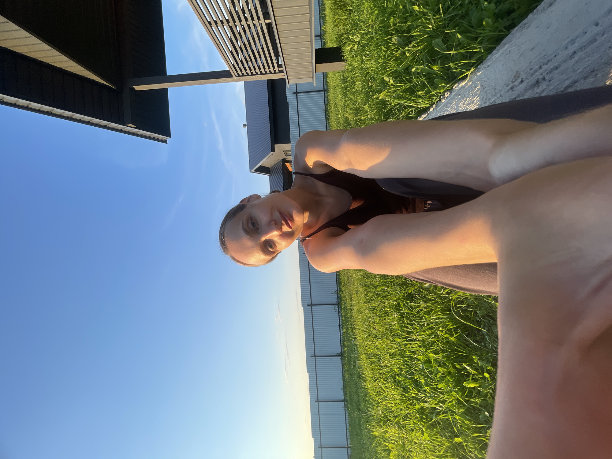 A young Caucasian woman is seated outdoors in a grassy area, possibly in Podshivalovo, Russia, with a clear blue sky above. She is wearing a dark tank top and dark pants. The camera is held by her, creating a selfie-style image where her arms and legs are prominently featured in the foreground, bathed in warm sunlight. Behind her, a white fence stretches across the landscape, and a dark-colored house with a modern design is visible, along with a covered patio area with horizontal slats. The grass is green and appears well-maintained. The lighting suggests it is either early morning or late afternoon.