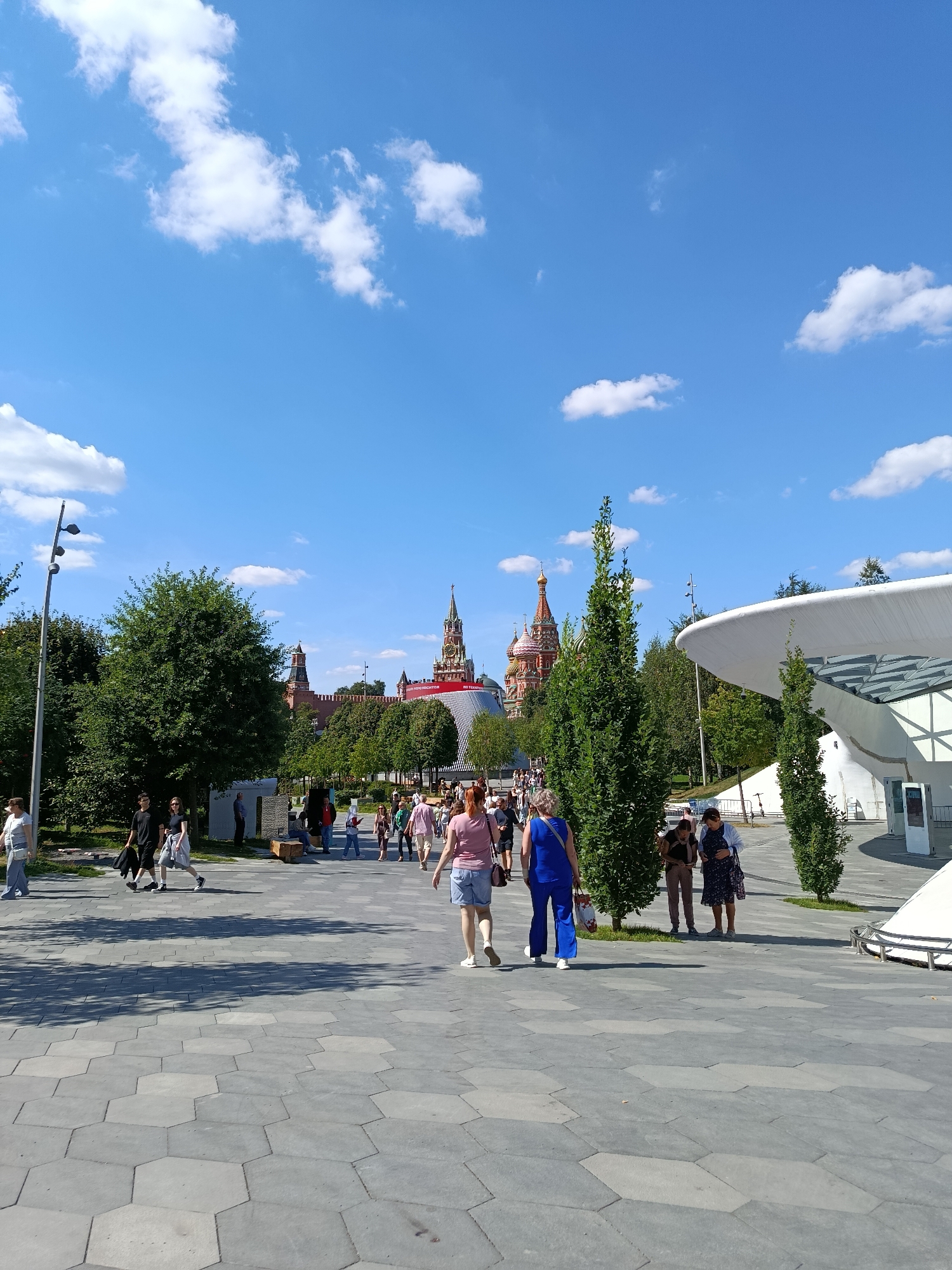 The image depicts a sunny day with a clear blue sky dotted with white clouds. A wide, hexagonal-tiled pathway leads through a park-like setting. In the background, several domed buildings, characteristic of Russian architecture, are visible, most notably St. Basil's Cathedral with its vibrant colors and distinctive onion domes. A modern, white, curved structure with glass panels is situated on the right side of the pathway. Numerous people are walking along the path, some singly, others in small groups, enjoying the outdoors. Trees with lush green foliage are scattered throughout the scene, casting shadows on the pathway. A lamppost with multiple lights stands on the left side of the image.
