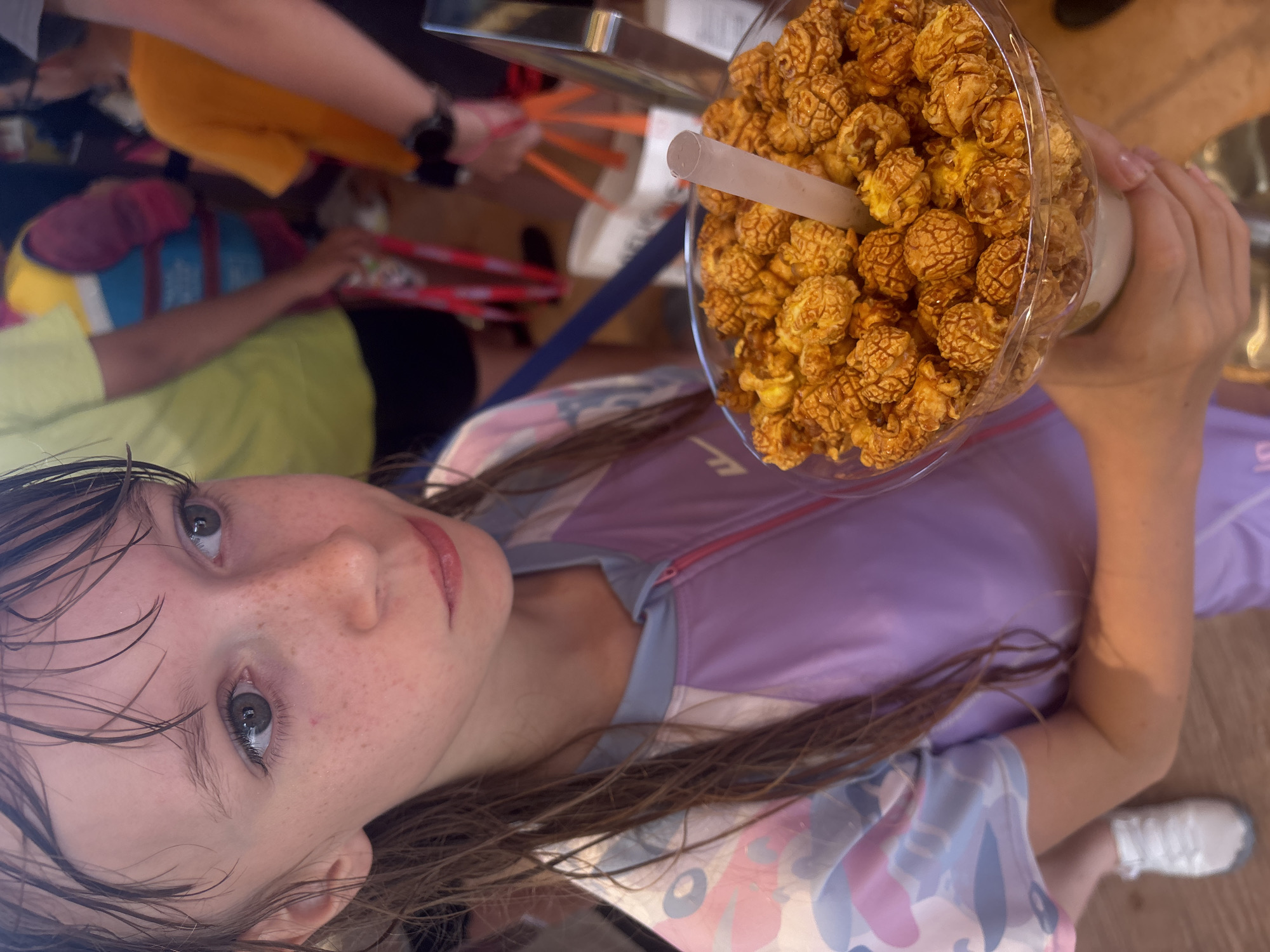 In this medium shot, a young girl is featured prominently, tilted with her face close to the camera, looking up and to the left. She has fair skin with freckles, light brown eyes, and dark brown hair that appears wet and is falling around her face. She is wearing a light purple and white zippered jacket with a pink zipper and a colorful pattern on the sleeves.
To the right of the girl, a large, clear plastic cup is filled with caramel popcorn. A white straw protrudes from the popcorn. A hand, likely belonging to an adult, is holding the cup. In the background, other people are visible, some holding red and orange objects that resemble chopsticks or straws. The setting appears to be outdoors, possibly at an event or a fair, given the festive atmosphere suggested by the background elements and the popcorn. The lighting suggests it is daytime. The girl's expression is one of curiosity and perhaps slight amusement. The scene is vibrant and full of life.