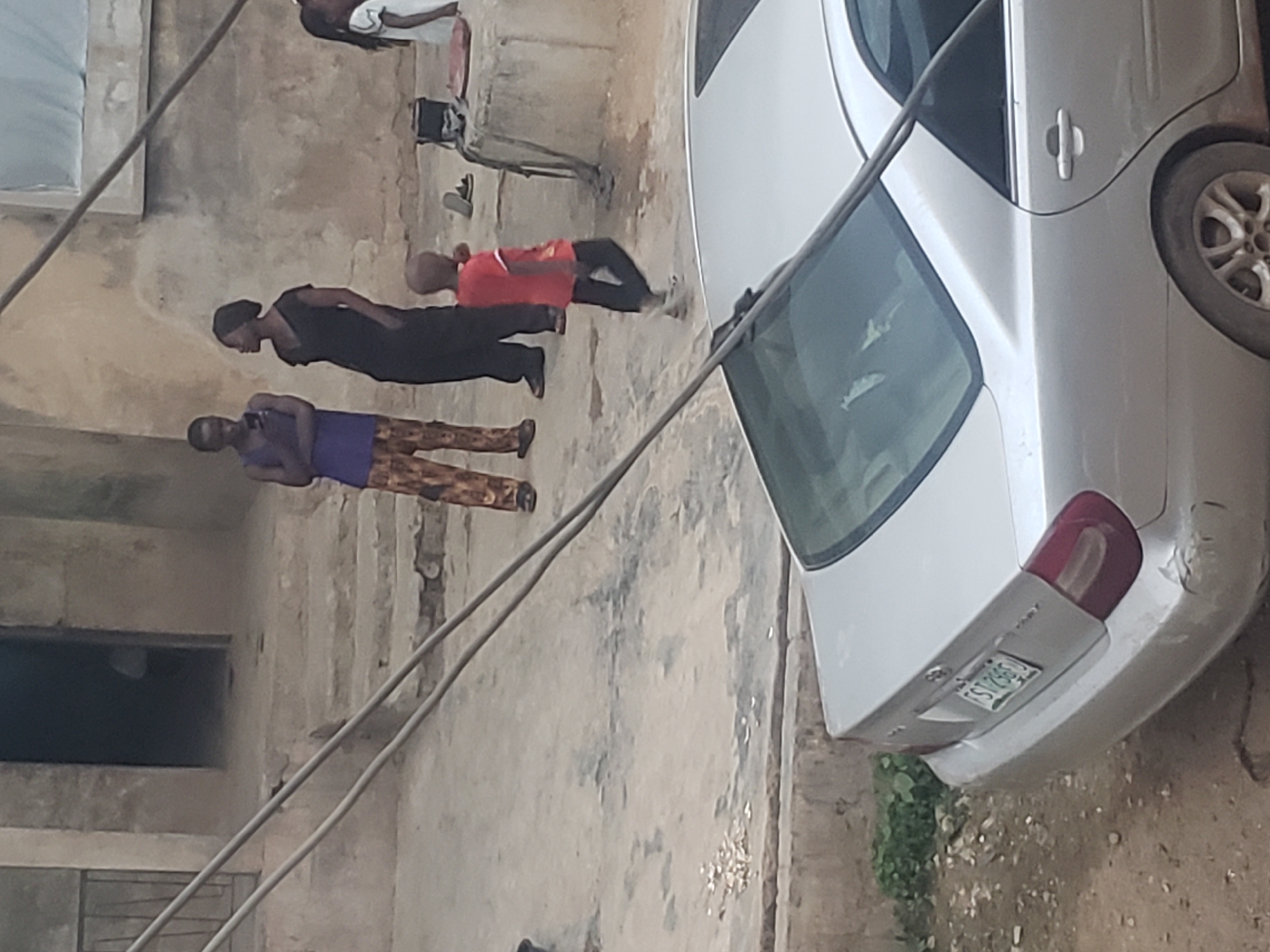 The image depicts a street scene in Alalubosa, Nigeria. The primary focus is a silver sedan parked on the right side of the frame. A group of four individuals are visible on the left side of the image, walking along a concrete walkway. The environment includes concrete building exteriors with weathered surfaces and a window. Power lines stretch across the scene, intersecting with the car.