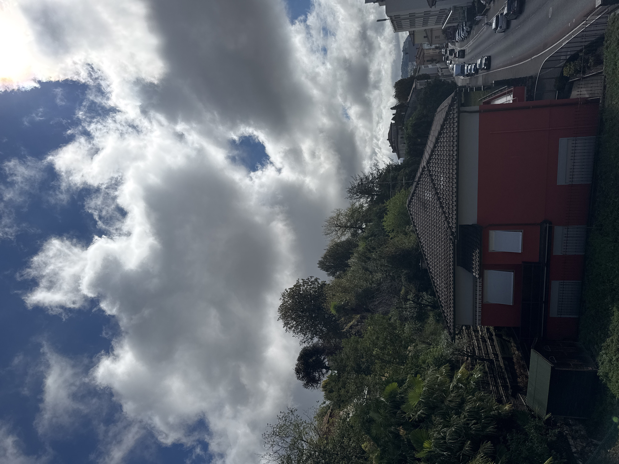 In Balerna, Switzerland, an image captures a cloudy sky with breaks of blue, casting dramatic shadows. The foreground features a vibrant red building with a dark-tiled roof, its windows covered by white blinds. To the left of the building, a small, dark green shed sits nestled amongst lush vegetation. Behind the red building, a hillside rises, densely populated with various trees and foliage. A winding road stretches into the distance on the right, lined with parked cars and a few vehicles in motion, suggesting an active urban or suburban setting. In the background, buildings and hills are visible, partially obscured by the clouds and distance.