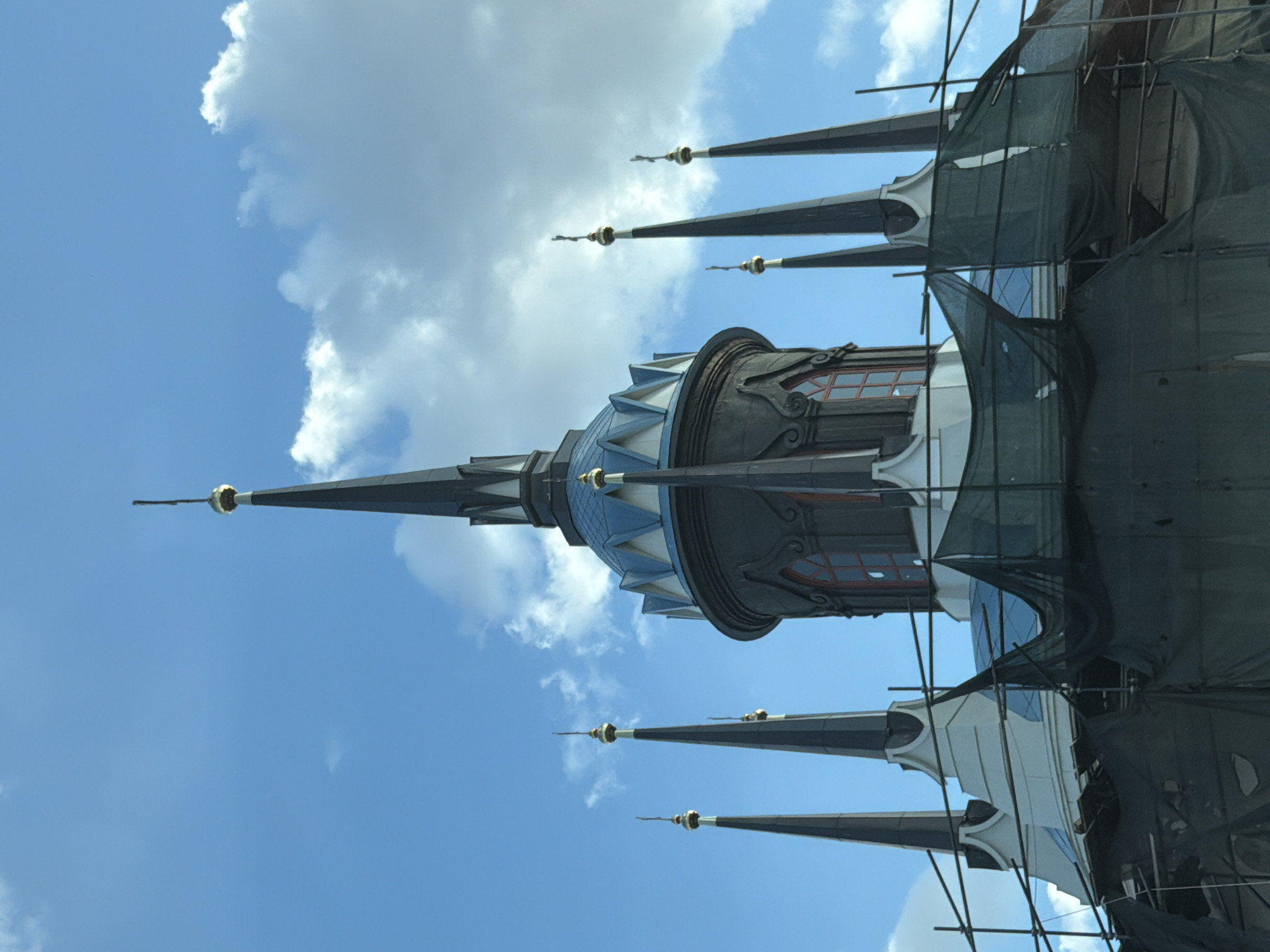 The image shows a low-angle view of the Church of the Vladimir Icon of the Mother of God in Bykovo, Russia. The camera points upwards towards the church's ornate spires and central dome, set against a background of a blue sky with scattered white clouds. Scaffolding and dark netting partially obscure sections of the church's exterior, indicating ongoing restoration or construction work. The church’s spires are dark and pointed, topped with gilded orbs. The architecture features intricate details and contrasting colors of blue, white, and dark grey.