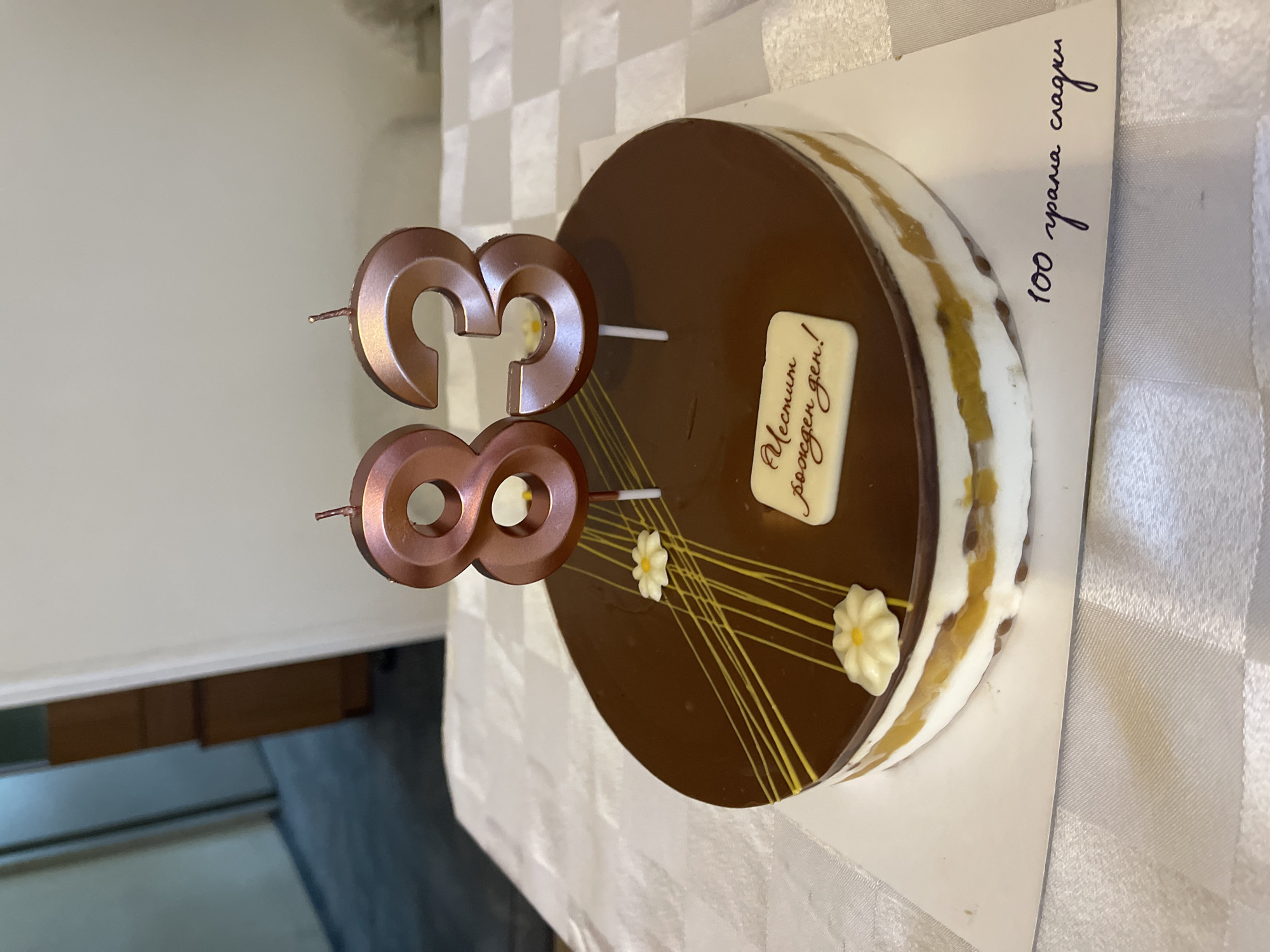 The image shows a chocolate birthday cake on a white checkered tablecloth, likely indoors. The cake is round with a dark chocolate glaze and a light-colored, multi-layered filling. Two candles shaped as the numbers "3" and "8" are placed on top, indicating the birthday is for someone turning 38. The cake is decorated with white icing flowers and thin yellow lines. A white rectangular plaque with brown writing in Cyrillic script that translates to "Happy Birthday!" is visible on the cake's surface. On the side of the cake board, more Cyrillic script reads "100 grams of sweetness". The overall setting is suggestive of a celebration, but no people are present in the frame. Based on the Cyrillic script, this image was likely taken in a region where the language is spoken, such as Bulgaria. If this picture was taken in Bulgaria, it is most likely taken in the capital city of Sofia.