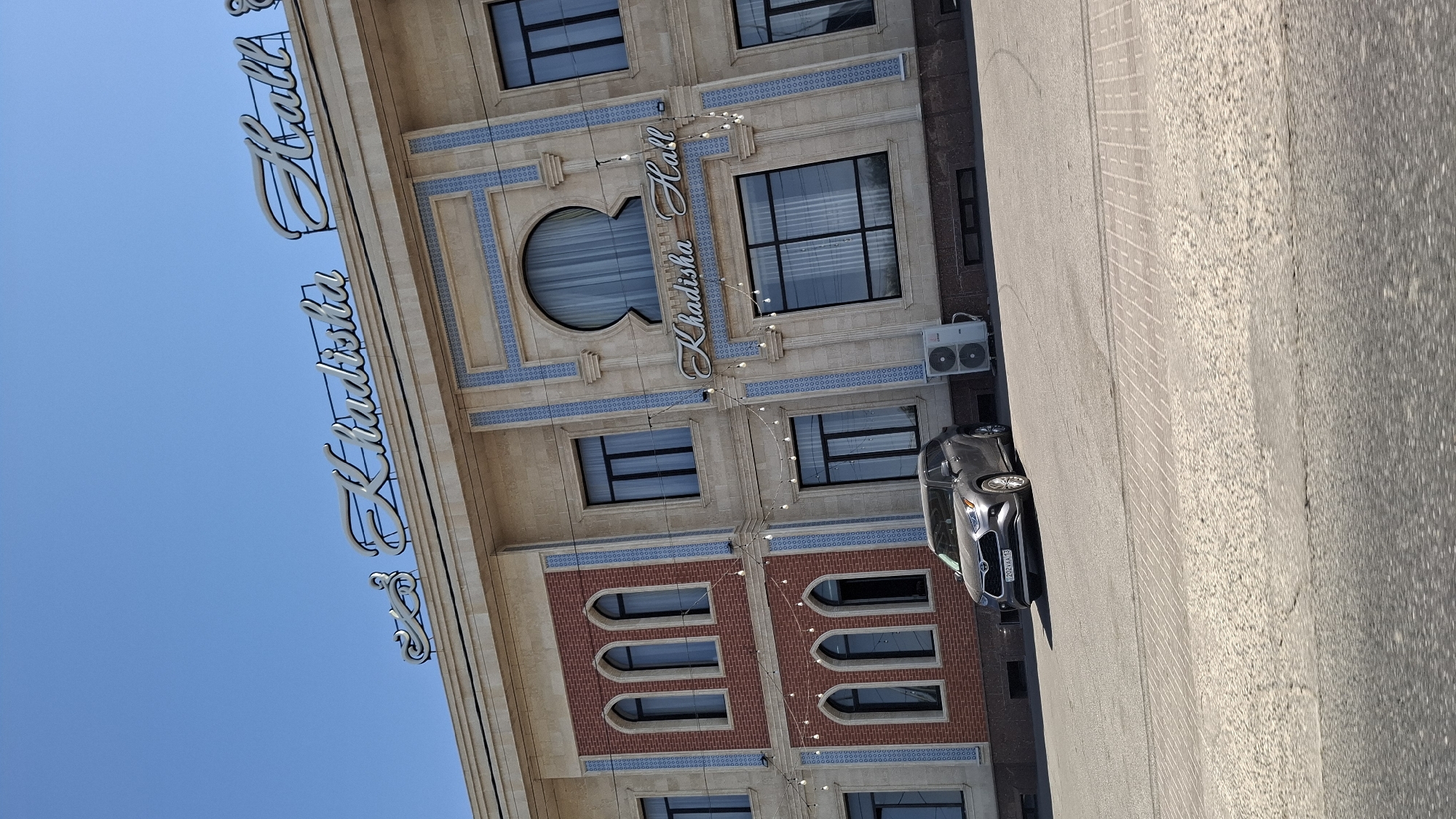 A mid-angle, exterior, eye-level shot shows a modern building with an ornate facade on a sunny day. The building has a tan-colored exterior with decorative blue tiling and red brick accents. The sign on the building reads "Khadisha Hall" in large, illuminated cursive letters. A silver SUV is parked in front of the building, and there are outdoor air conditioning units visible. The sky is clear and blue. The scene is located in the city of Shymkent, Kazakhstan.