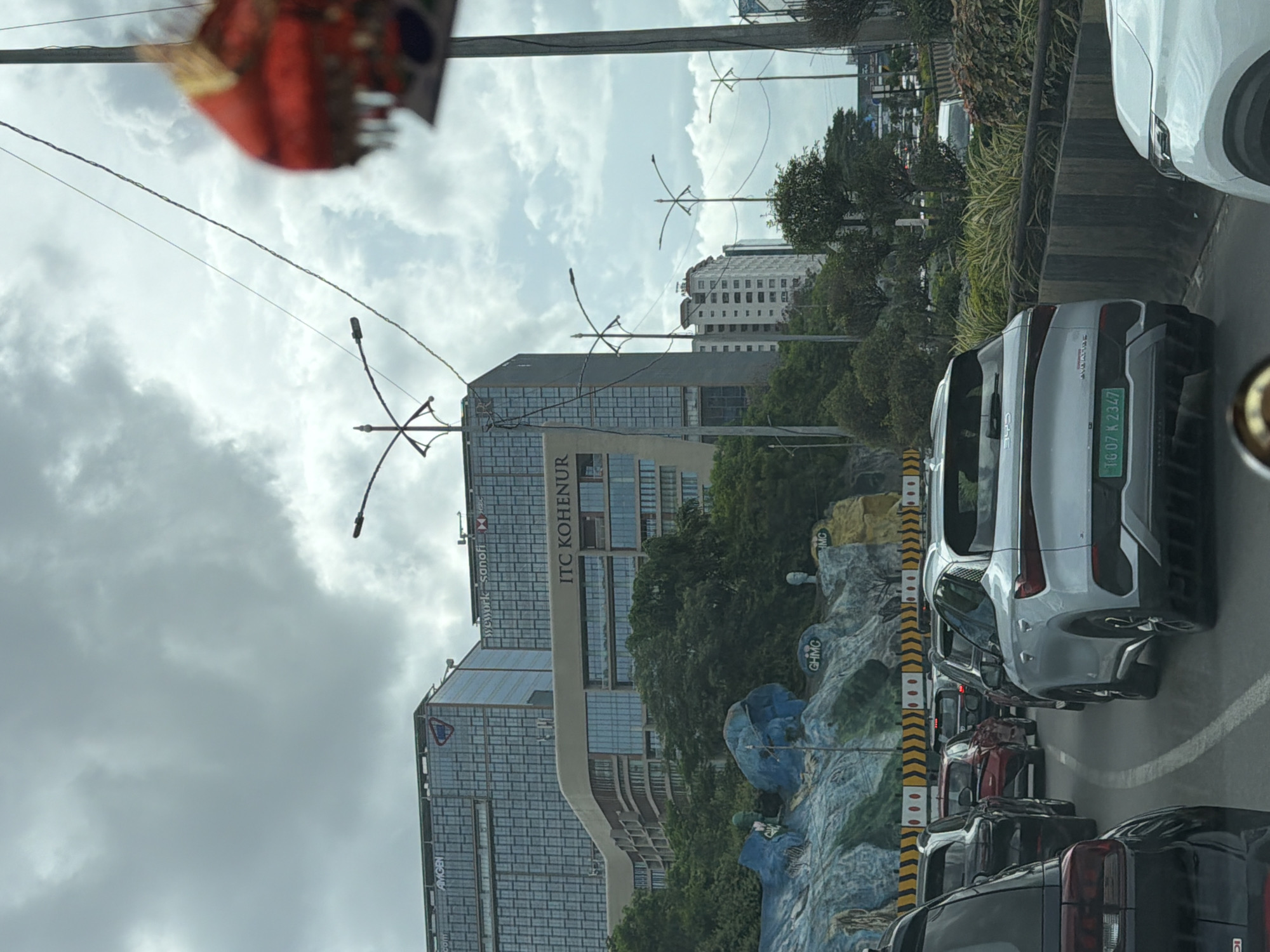 The image depicts a traffic jam on a cloudy day in Hyderabad, India, specifically on the Outer Ring Road near the ITC Kohenur hotel. In the foreground, a silver BYD Atto 3 electric car is prominent, displaying the license plate "TG 07 K 2347." Behind it, other vehicles like a black SUV and a red car are visible. The background features a section of the ITC Kohenur building, which is a large hotel, and other office buildings with signage such as "Amgen" and "WeWork Sanofi HSBC." A distinctive rock formation with painted murals of animals and human heads is also part of the landscape. The scene is characterized by heavy traffic, overhead electrical wires, and streetlights, indicating an urban environment.
