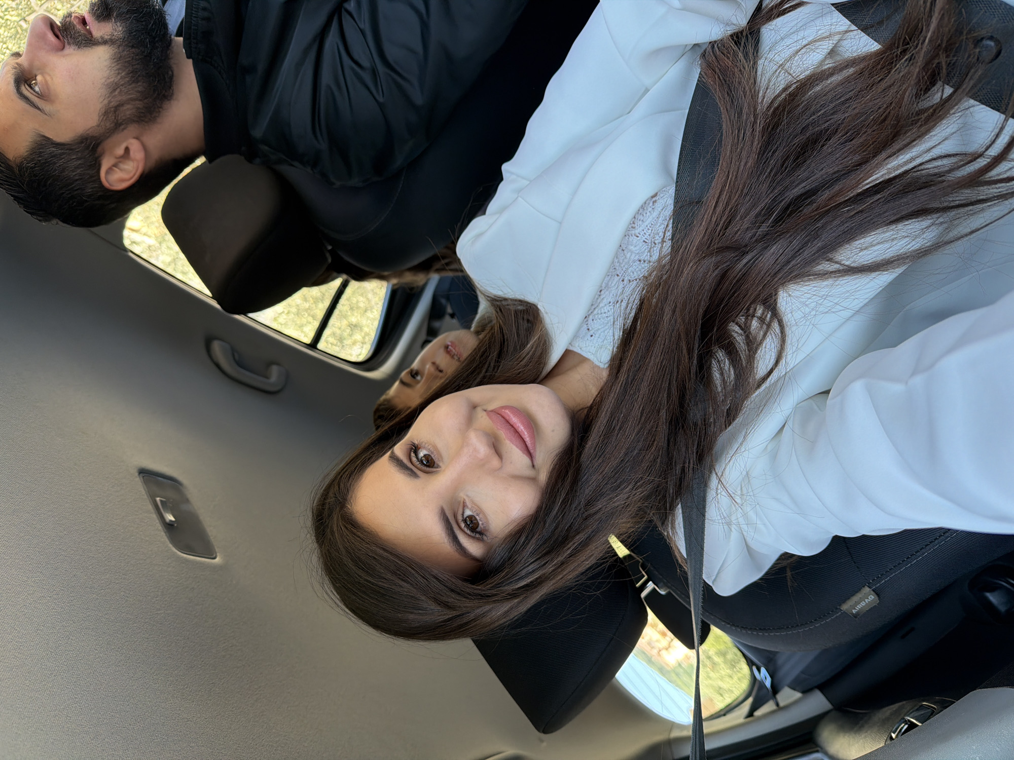 A young woman with long, dark brown hair is the primary subject of this selfie-style image, taken from inside a car. She is seated in the front passenger seat, looking directly at the camera with a soft, pleasant smile. Her brown eyes are open, and she wears pinkish-nude lipstick. She is dressed in a white top, possibly a blazer or blouse, with a subtle lace pattern visible on the chest. A dark seatbelt crosses her torso.
In a reflection visible behind her, likely from a rearview mirror or a reflective surface within the car, a man with dark hair and a beard can be seen. He is wearing a black jacket and appears to be in the back seat.
The car's interior is contemporary, featuring a light grey headliner and dark fabric headrests. The word "AIRBAG" is visible on the side of the passenger seat. Bright daylight streams through the car windows, revealing glimpses of green foliage outside, suggesting the car is outdoors during the day in good weather. The scene implies the two individuals are traveling together, possibly in Budva, Montenegro.