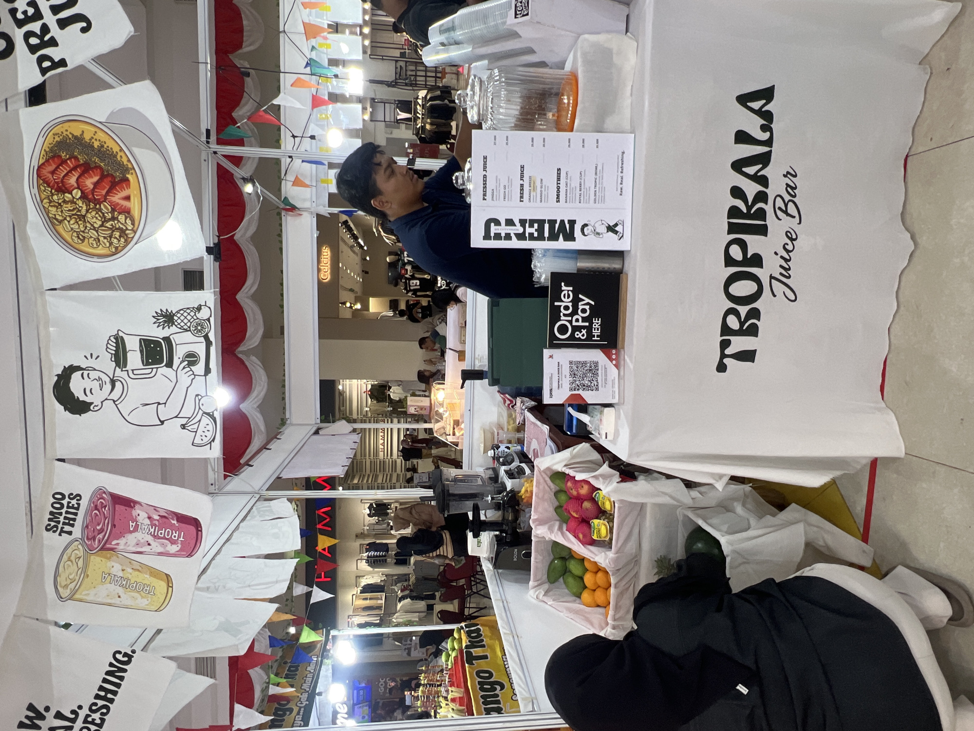 The image shows a juice bar stall at what appears to be a market or festival in Banyurojo, Indonesia. The stall, named "TROPICALA Juice Bar," features a white tablecloth printed with its logo. In front of the stall, a man in a dark blue shirt is seen behind a counter, preparing drinks. On the counter are several items, including a blender, a juicer, containers of fruit like oranges and apples, and a menu board listing various juice and smoothie options. A "Order & Pay Here" sign is also visible. In the background, there are displays of clothing and other market stalls, with various decorative flags and banners hanging overhead. Two people are partially visible in the foreground, one wearing a black hijab.
