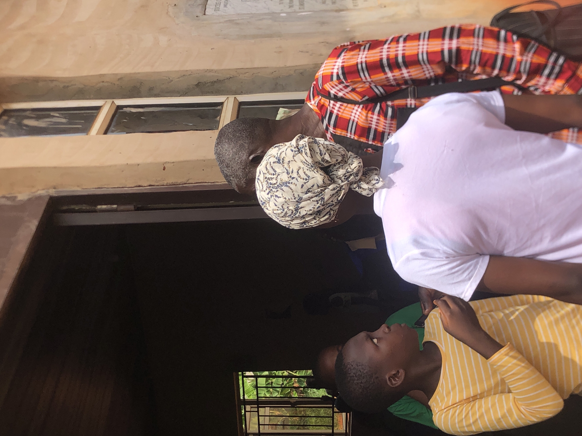 Four individuals are positioned near the entrance of a building. A child in a yellow striped shirt is looking upwards. A person wearing a white shirt is standing nearby. An older man with a patterned head covering and a red and orange plaid shirt is also present. The building has a dark interior and a barred window. The location is Ukwu-Abwa, Nigeria.