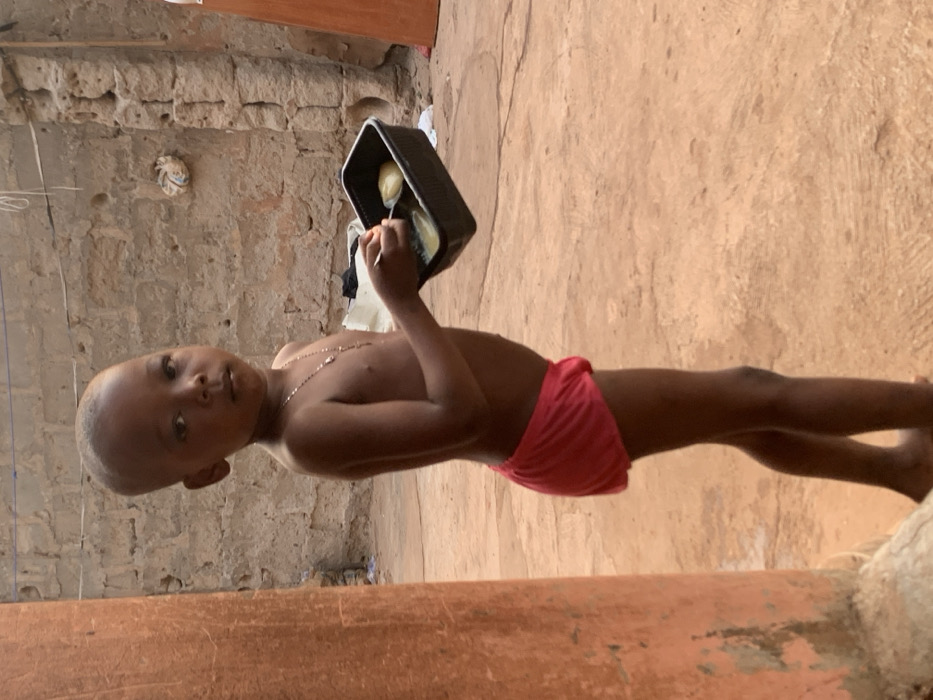 A young, shirtless child with close-cropped hair wears red shorts and a silver necklace. He stands on a brown, textured surface, holding a black container with food and a spoon. The child looks towards the camera. Behind him, a wall constructed of rough, tan-colored blocks is visible. In the background, a light-colored cloth item hangs. A weathered red-brown structure is in the foreground and partially obscures the lower portion of the image.