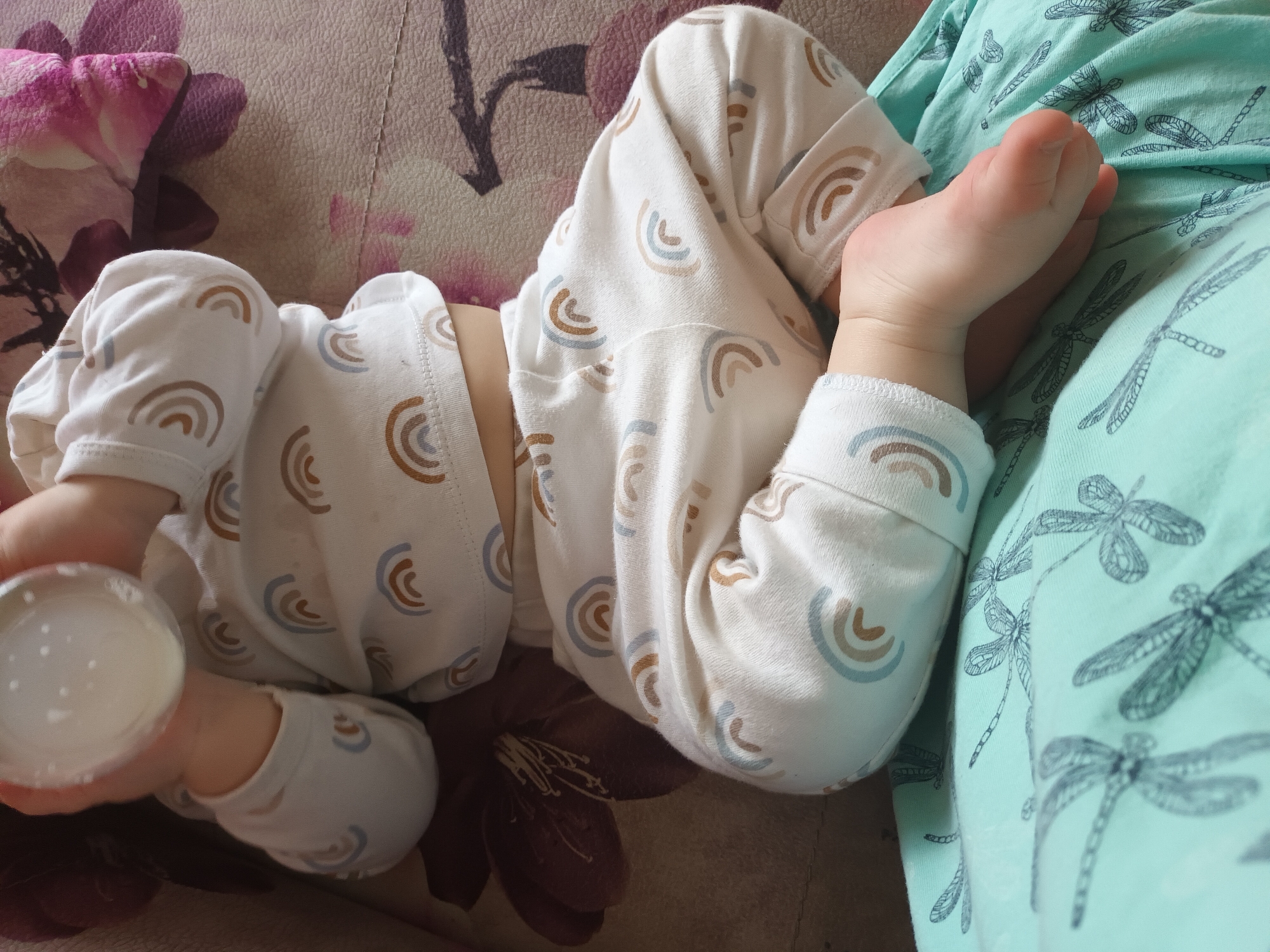 A baby wearing white pajamas with a rainbow pattern lies on a patterned surface, possibly a couch or bed. The baby is holding a small, round container in its left hand. Part of the baby's body is resting on a light green fabric with a dragonfly pattern. The location is Ольховка, Russia.