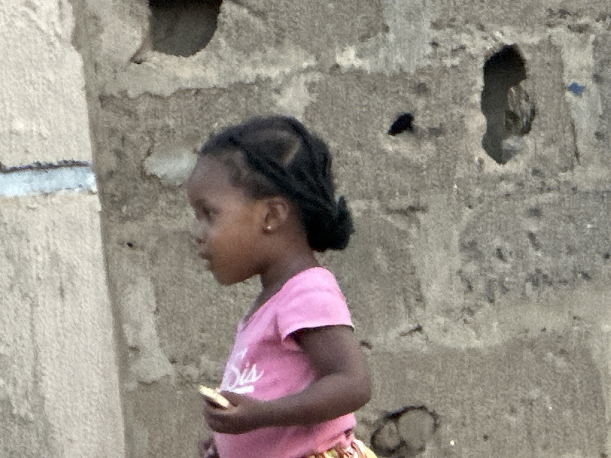 A young, dark-skinned girl stands in profile, holding a small, light-colored object in her left hand. She wears a pink short-sleeved shirt and has her hair styled in braids. The background is a rough, textured wall with a few holes or indentations. The location is Unknown, Unknown.